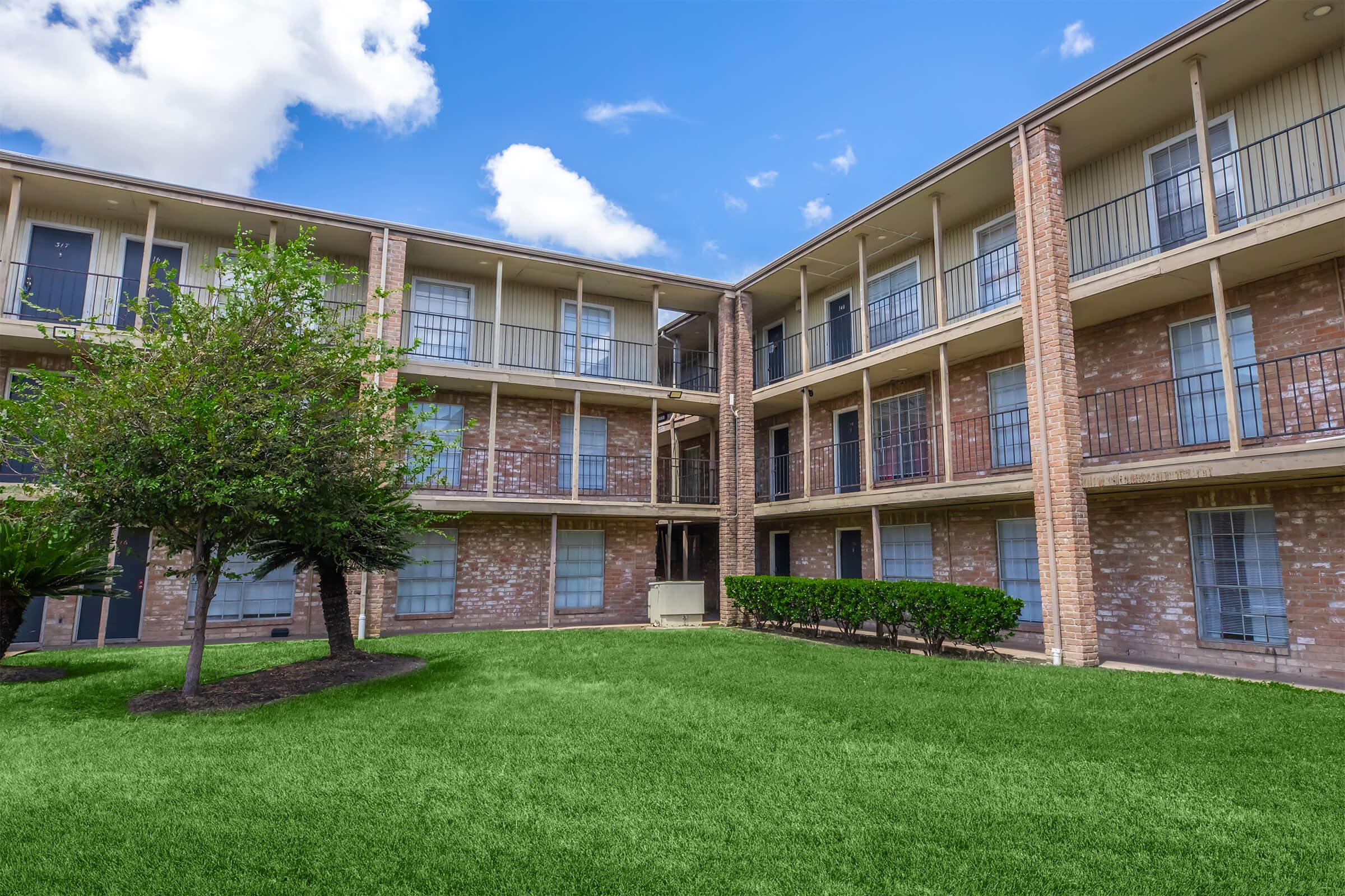 A view of an apartment complex featuring three levels of balconies surrounded by greenery. A small tree is visible in the courtyard area, with well-maintained grass and a blue sky with scattered clouds in the background.