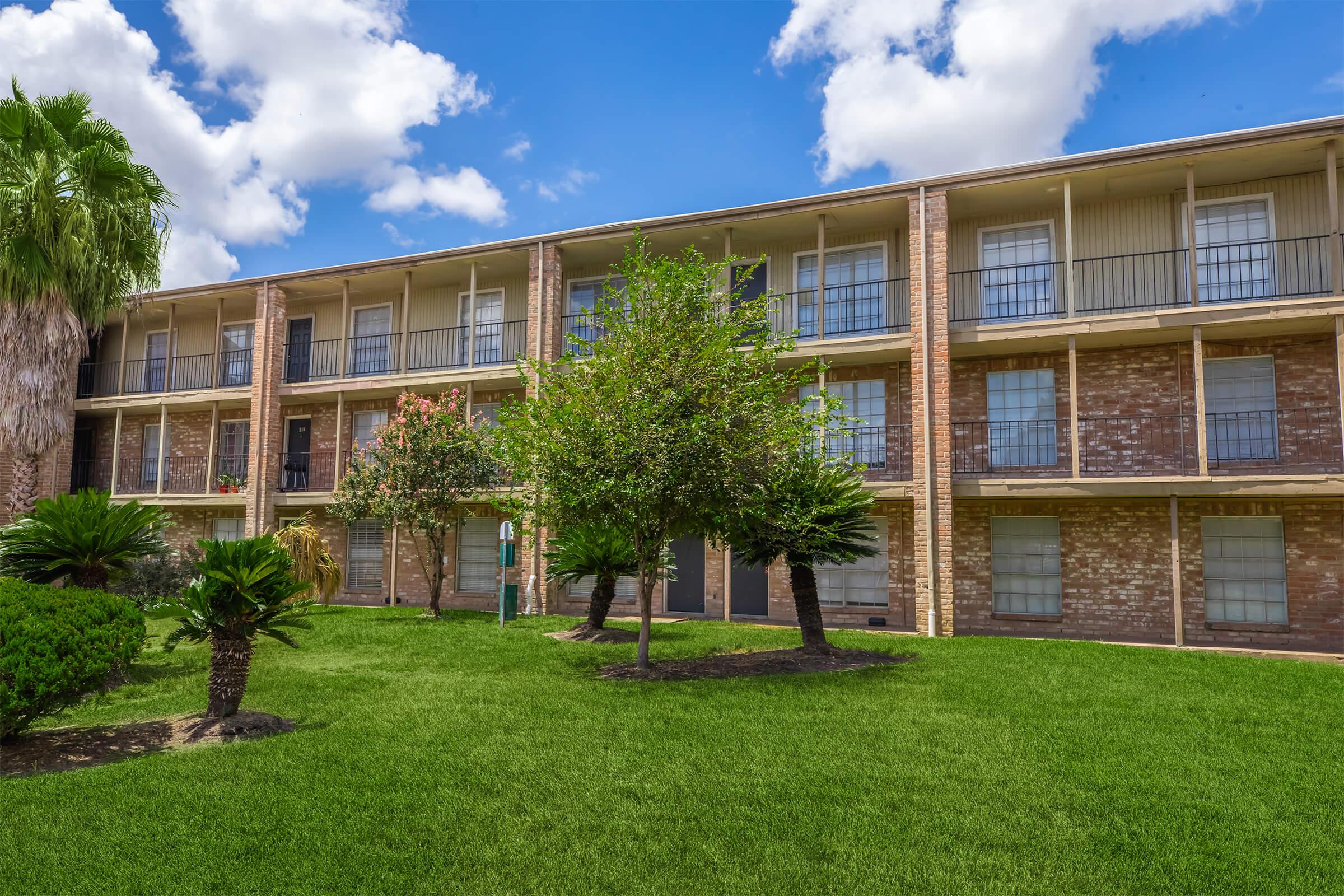 Three-story brick apartment building with multiple balconies. Surrounding greenery includes palm trees and shrubs. The sky is partly cloudy, with blue patches visible. The lawn is well-maintained and features lush grass.