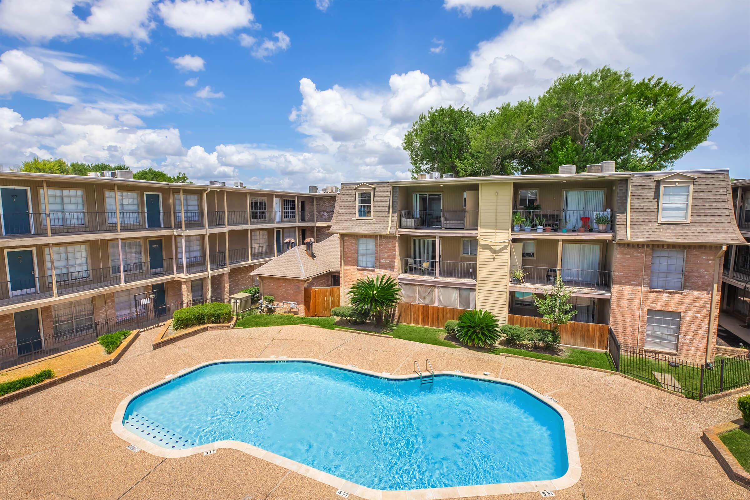 A view of an apartment complex featuring multiple buildings surrounding a swimming pool. The pool is oval-shaped and appears inviting, surrounded by landscaped greenery. Fluffy clouds and blue skies create a bright and sunny atmosphere, enhancing the appeal of the outdoor space.