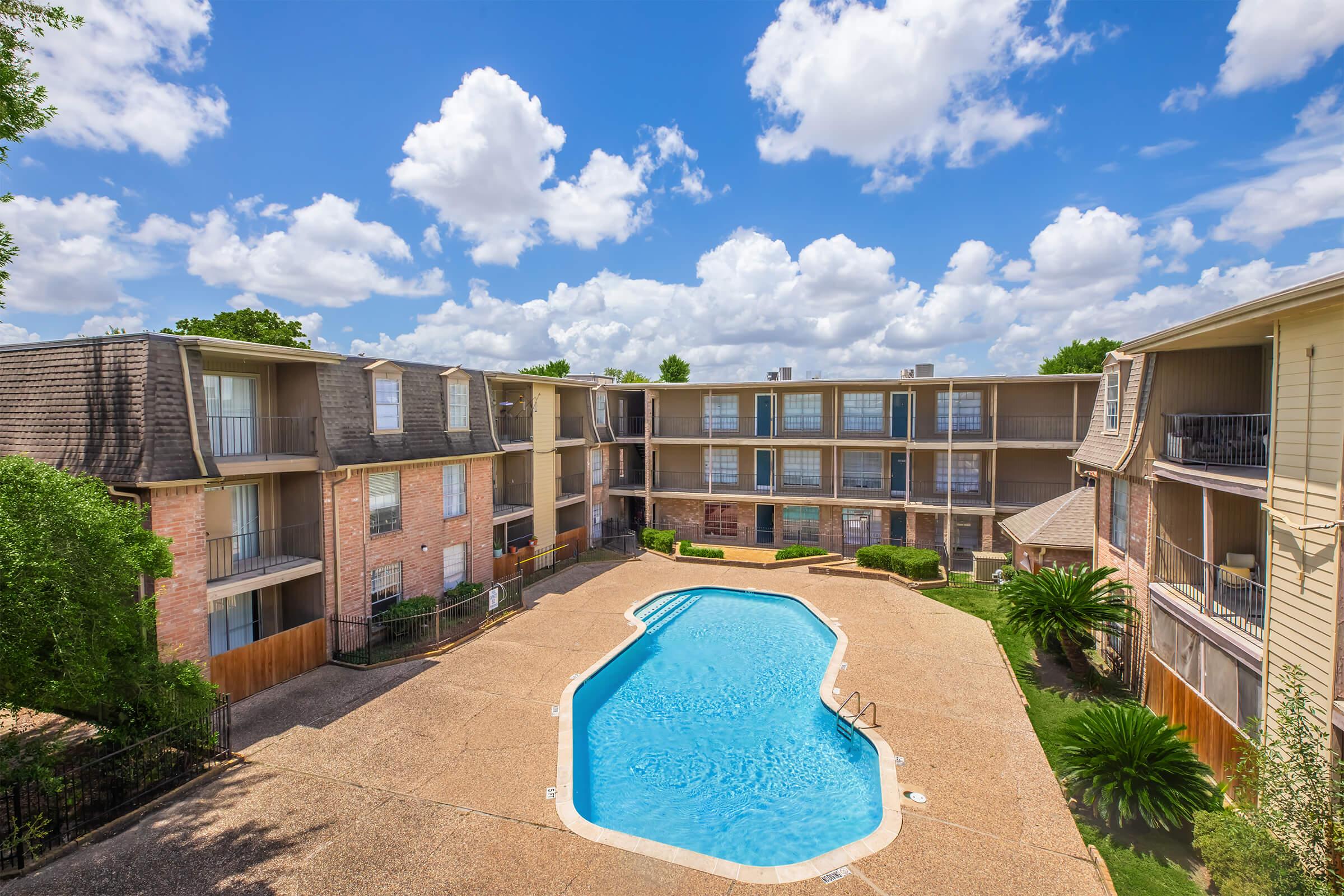 A well-maintained apartment complex featuring a central swimming pool surrounded by landscaped greenery. The building boasts multiple floors with balconies, and the sky is partly cloudy, creating a bright and inviting atmosphere.