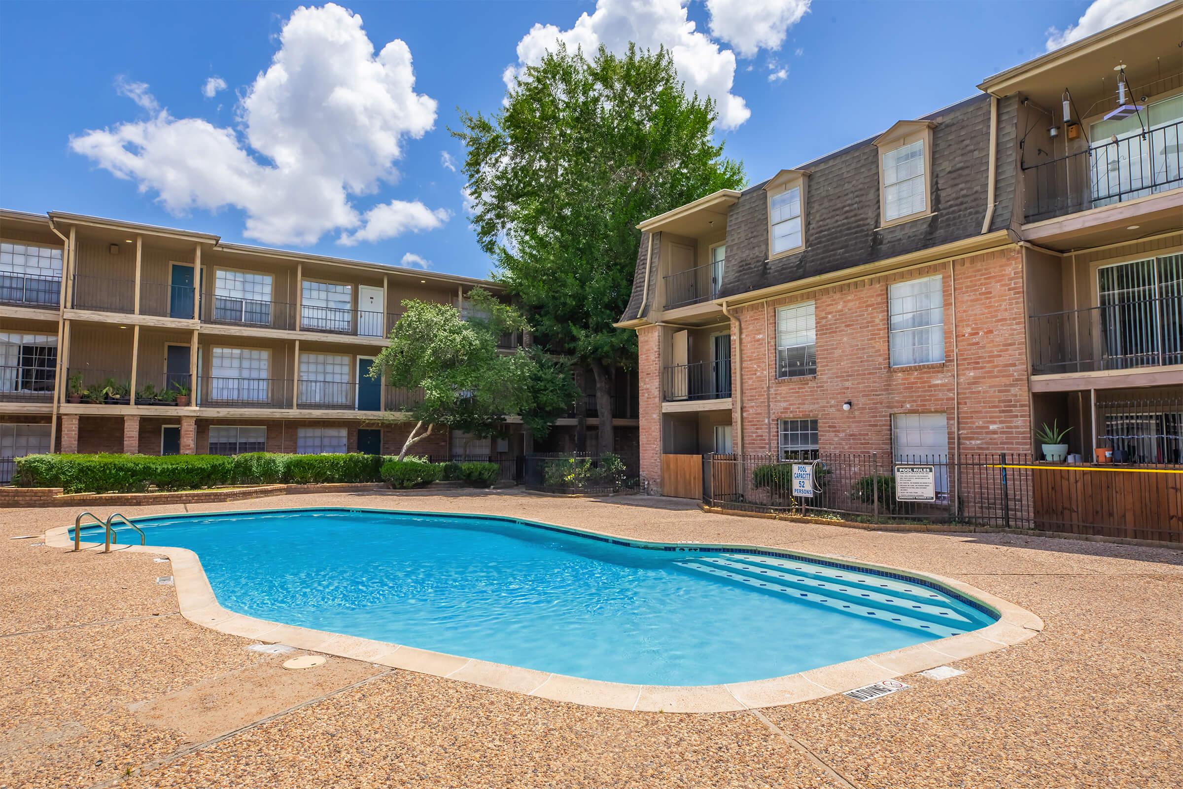 A clear outdoor swimming pool surrounded by a landscaped area, with two residential buildings in the background. The pool features a gradual entry and is bordered by textured concrete. Lush green trees provide shade, and a blue sky with scattered clouds is visible above.