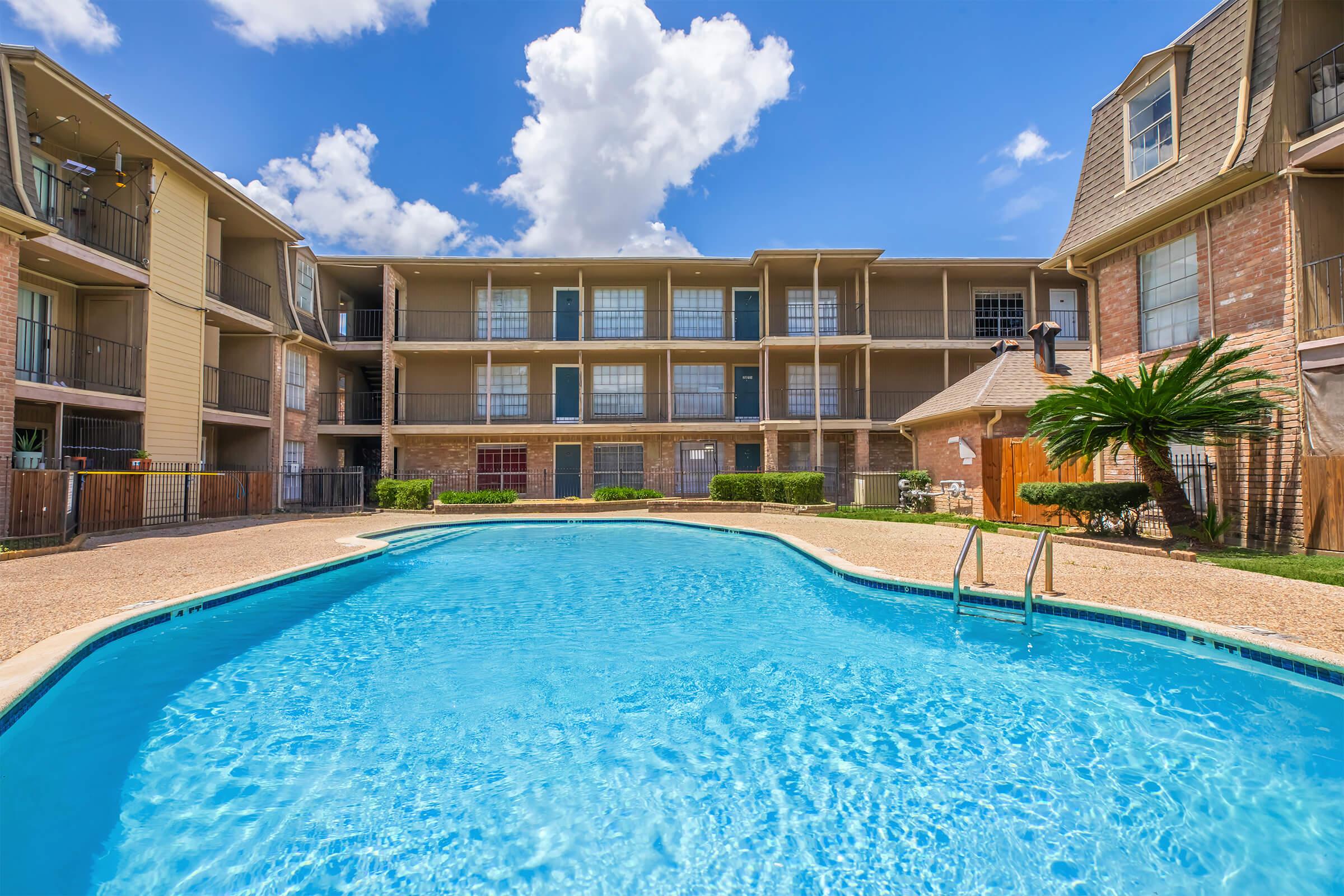 A clear blue swimming pool in the foreground, with a surrounding area featuring lush green grass and palm trees. In the background, there are three multi-story apartment buildings with balconies, under a bright blue sky filled with white, fluffy clouds.