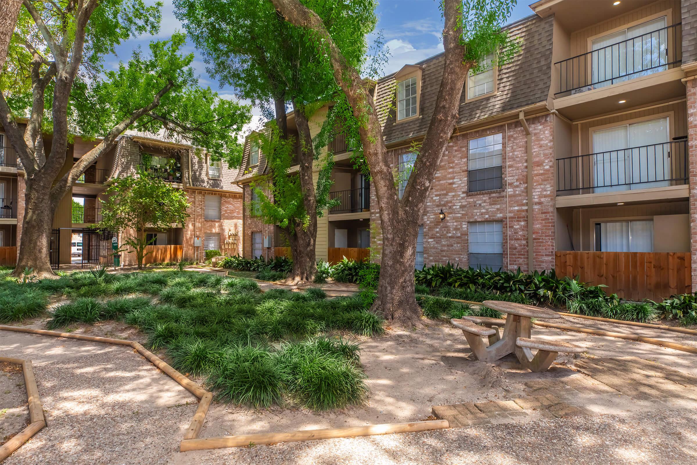 A landscaped courtyard featuring several trees and shrubs, surrounded by brick apartment buildings. Wooden benches are placed on a gravel path, providing a peaceful outdoor space for residents. The scene is bright and inviting, with a clear blue sky visible above the buildings.