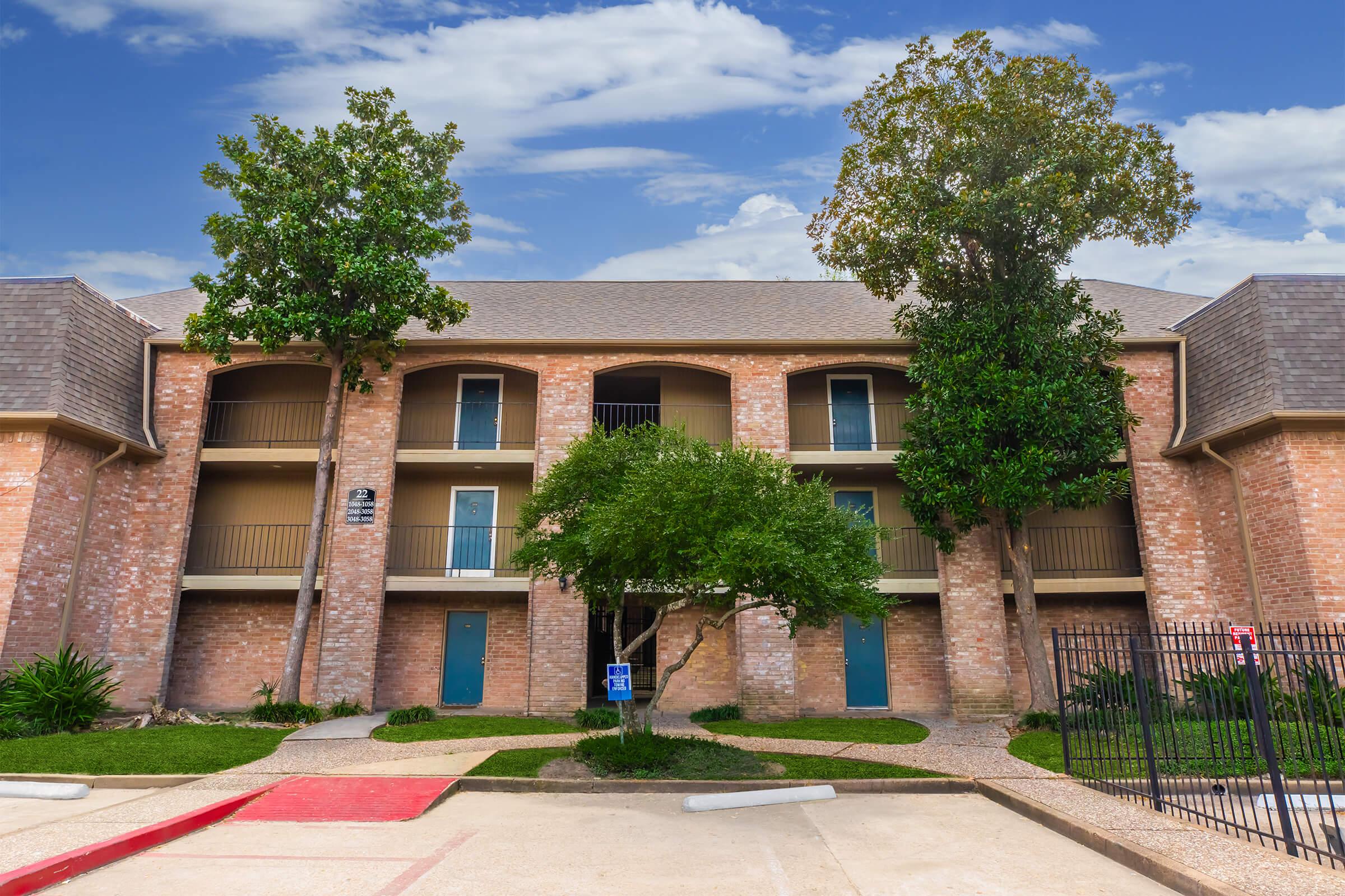 A two-story apartment building with brick exterior, featuring balconies on each level. Lush green trees are planted on either side of the entrance, which has a small grassy area. The sky above is partly cloudy, creating a bright and inviting atmosphere. A parking area is visible in front.