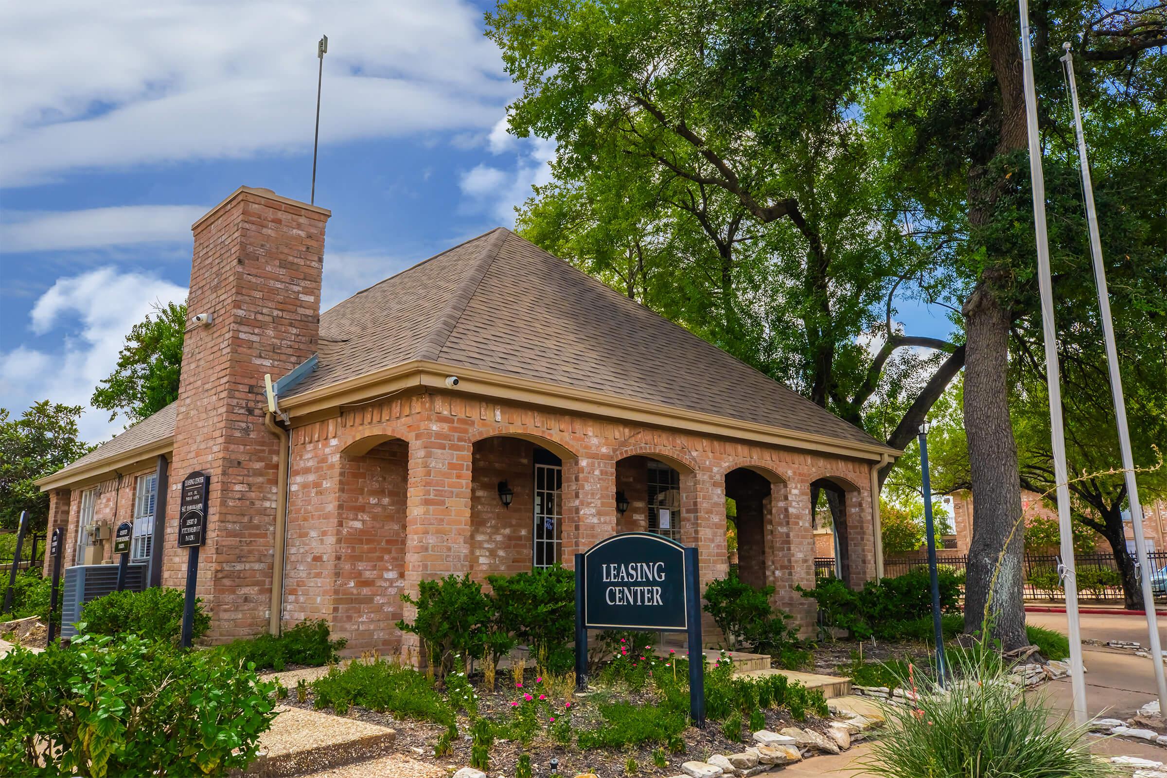 A brick building with a pitched roof and large arched windows, surrounded by trees and landscaping. A sign in front reads "Leasing Center," indicating it is likely an office for a residential or commercial property. The sky is partly cloudy, suggesting a pleasant day.