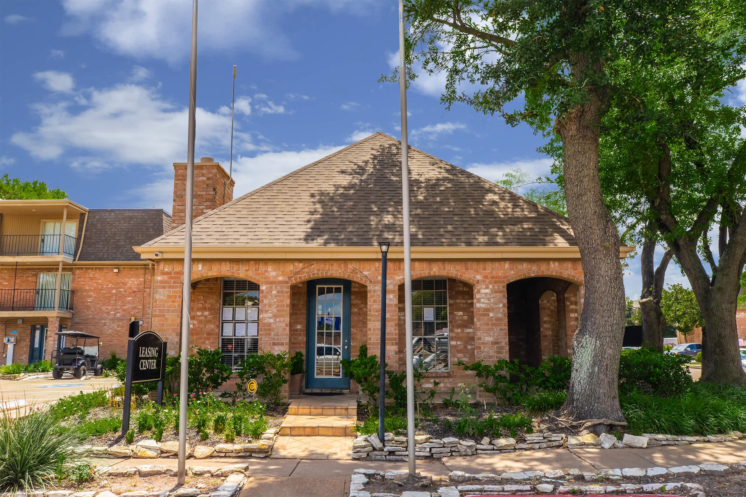 A brick building with a sloped roof, featuring a central blue door and large windows. Surrounding the structure are well-maintained gardens and trees. Two flagpoles are positioned in front of the building, and there is a pathway leading up to the entrance. The sky is clear with a few clouds.