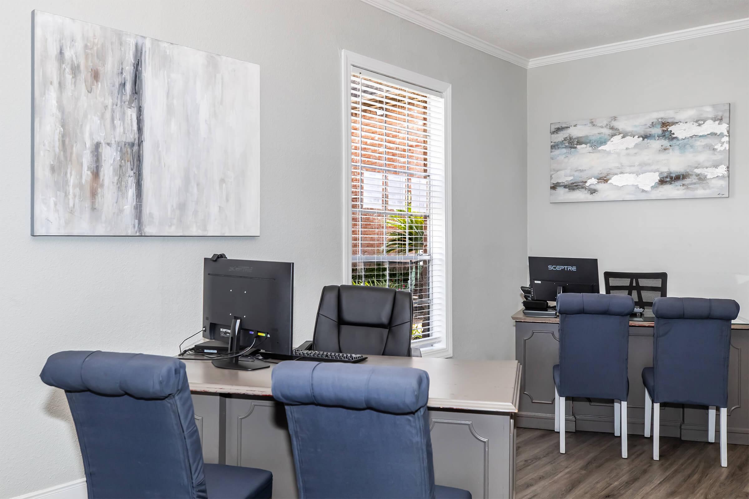 A modern office space featuring two workstations with computers, each accompanied by navy blue upholstered chairs. The walls are adorned with abstract artwork, and natural light filters in through a window, illuminating the clean and organized environment.