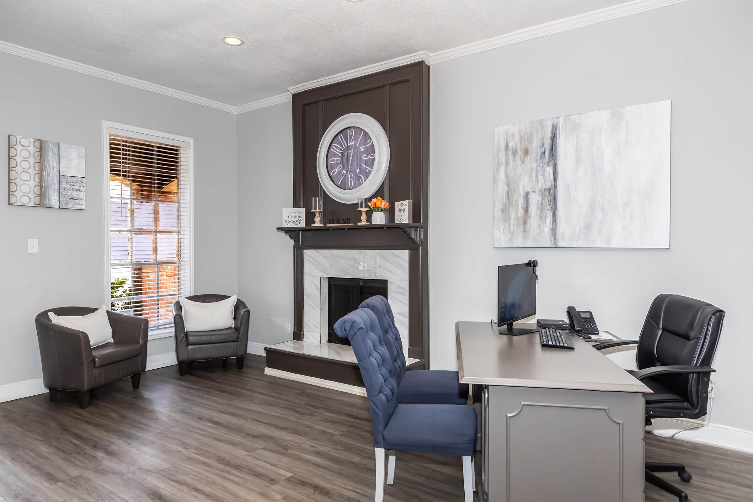 A modern office space featuring a desk with a computer and phone, a fireplace with a large clock above it, and two armchairs. The walls are painted in light gray, and there are decorative wall art pieces. Natural light comes in through a window with blinds, creating a welcoming atmosphere.