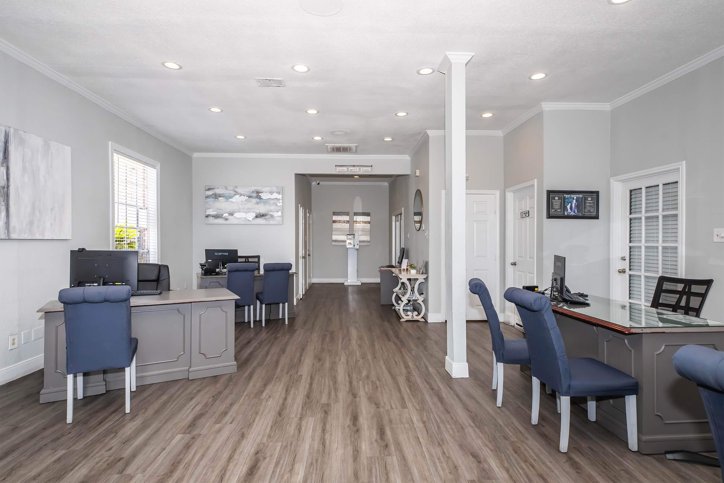 A modern office reception area featuring two desks with computers, blue upholstered chairs, and wood-like flooring. The walls are painted light gray, and there is artwork and a mirror on display. Bright lighting enhances the spacious feel of the room, creating a welcoming atmosphere.