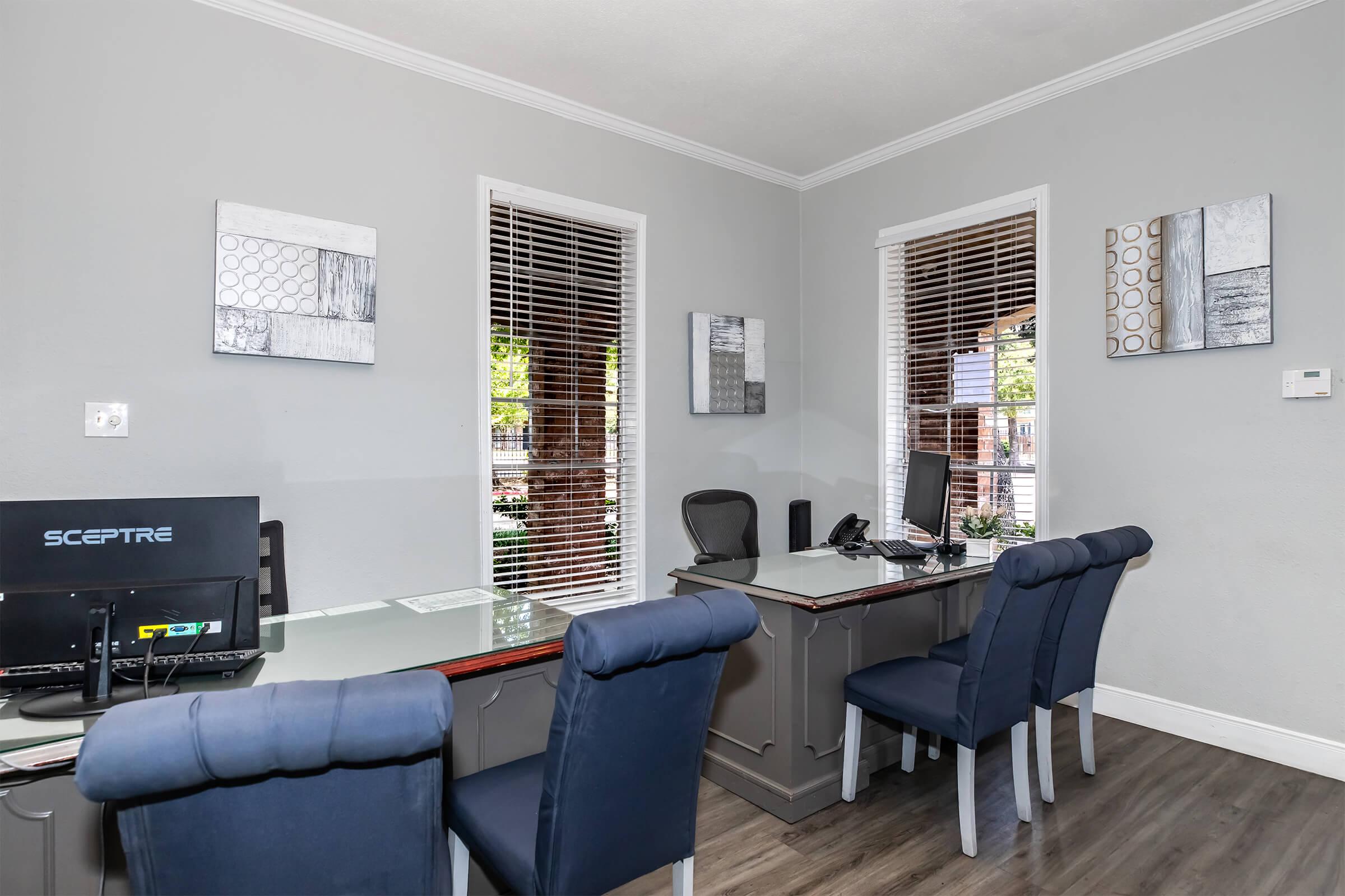 A modern office space with a large desk, computer monitor, and black office chair. Two blue upholstered chairs are positioned around a light-colored table. The walls are painted gray and feature abstract artwork. Window blinds allow natural light to enter the room, creating a bright and inviting atmosphere.