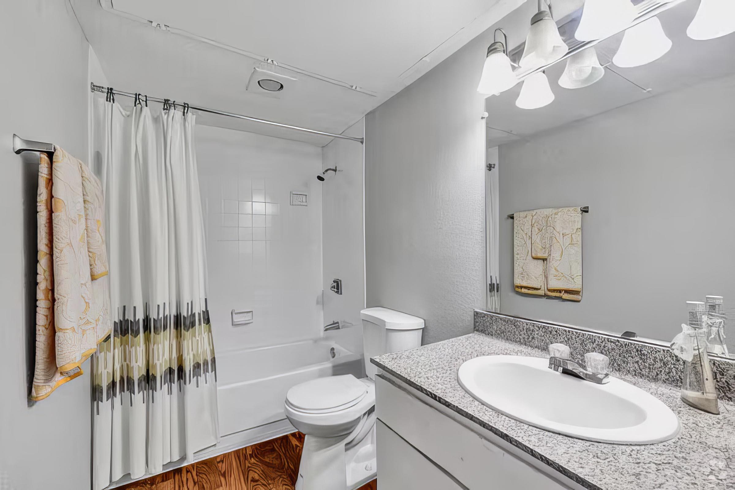 A modern bathroom featuring a white bathtub with a shower curtain, a white toilet, a large mirror above a granite countertop with a sink, and under-cabinet lighting. The walls are painted gray, and a towel hangs near the sink. The floor has wooden planks, adding warmth to the space.