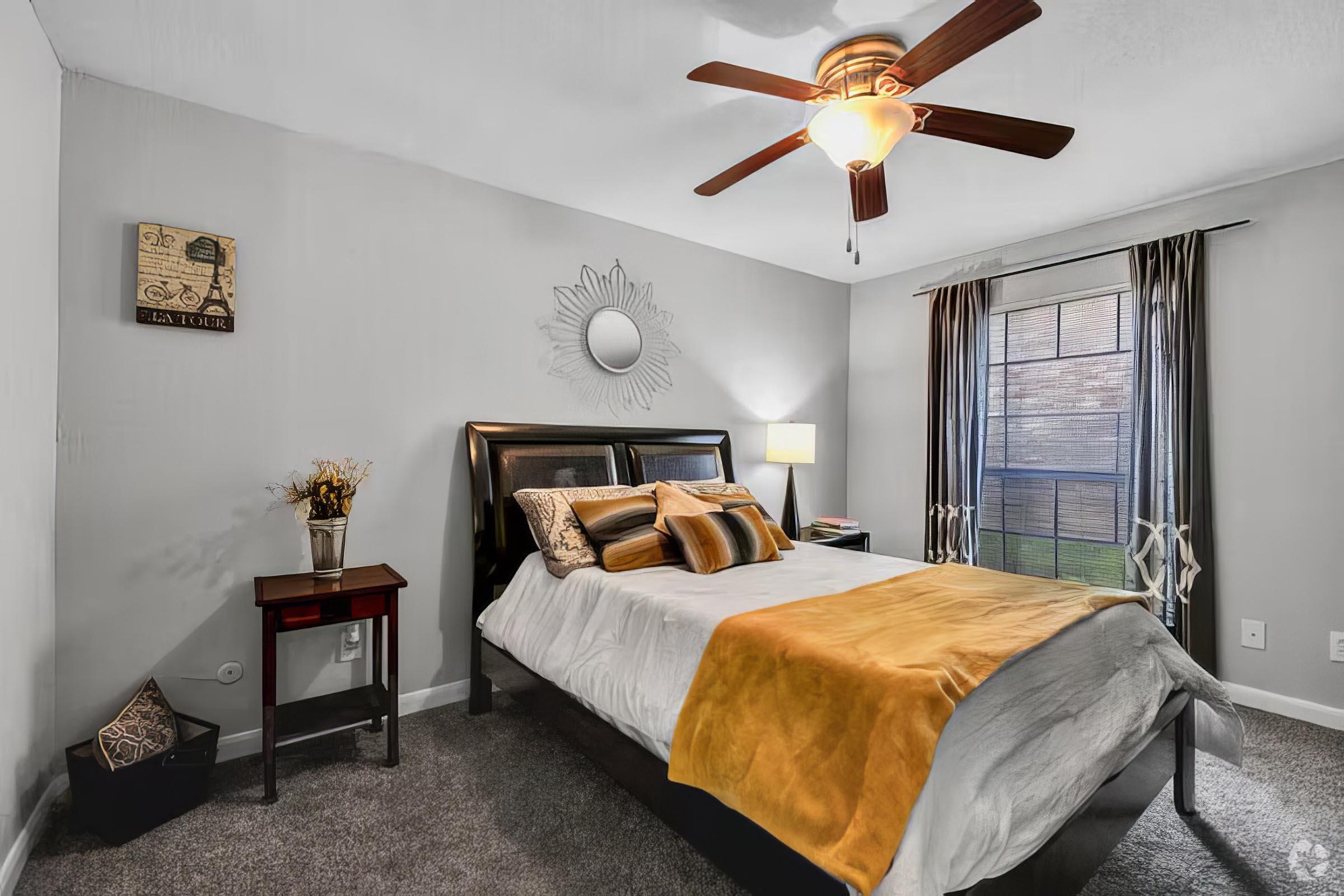 A cozy bedroom featuring a bed with a white and yellow comforter, a ceiling fan with wooden blades, and a decorative mirror on the wall. The room has a window with light-filtering curtains, a bedside lamp, and a small decorative plant. The walls are painted in a soft gray color, creating a calm atmosphere.