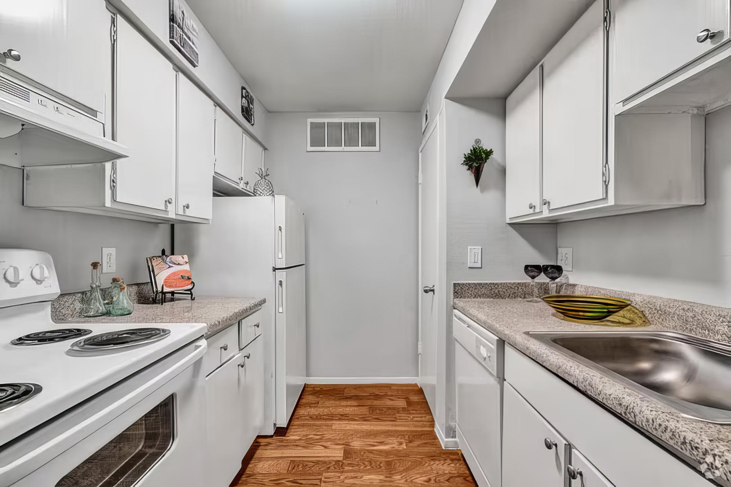 A bright, modern kitchen featuring white cabinetry, a countertop with a sink, and an electric stove. The walls are painted light gray, and there is a refrigerator visible. Decorative items include a small plant and a framed picture. The hardwood floor adds warmth to the space.