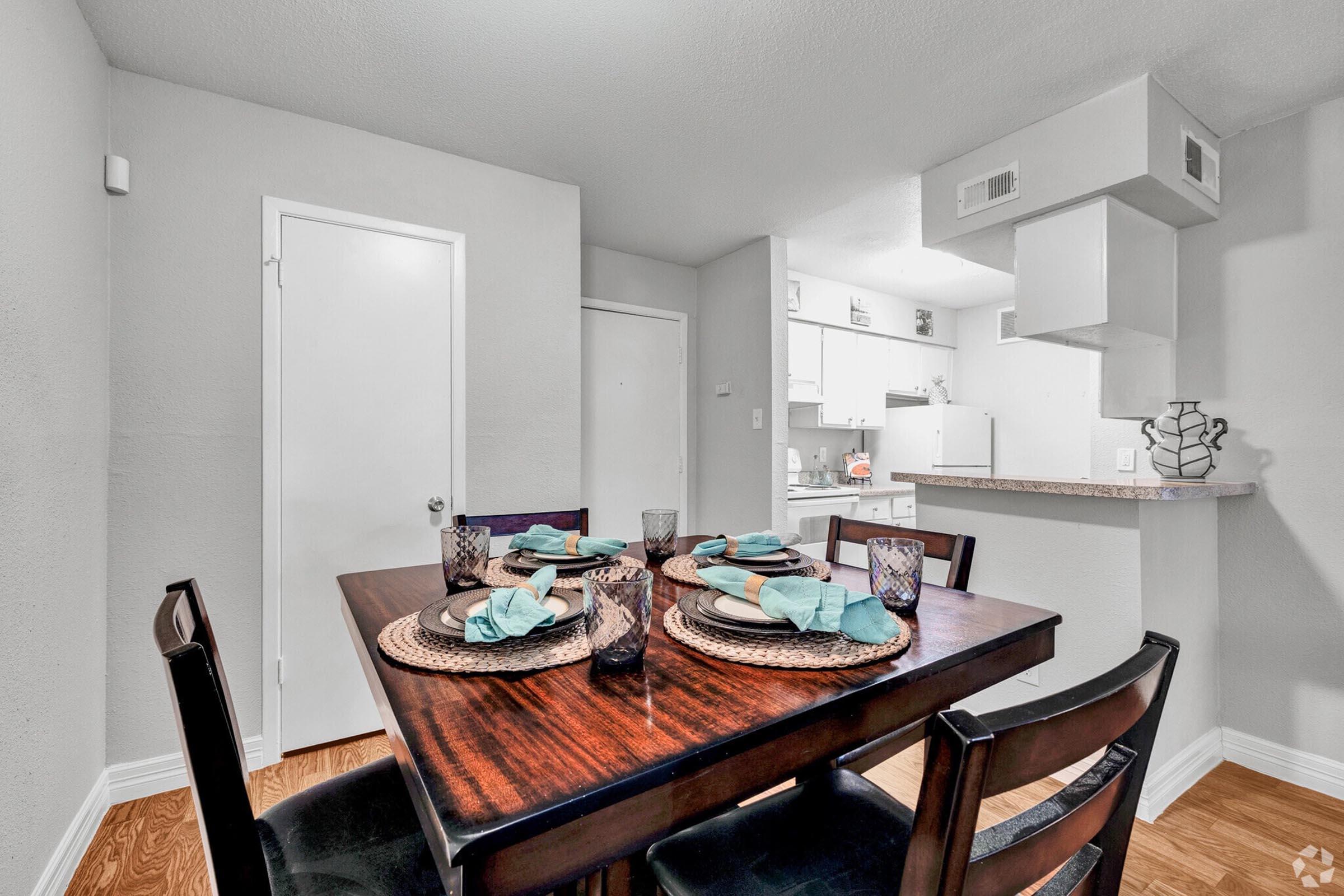 A dining table set for four, featuring turquoise napkins, woven placemats, and decorative glasses. The background shows a modern kitchen with white cabinets and a light-colored wall, creating a cozy and inviting atmosphere.