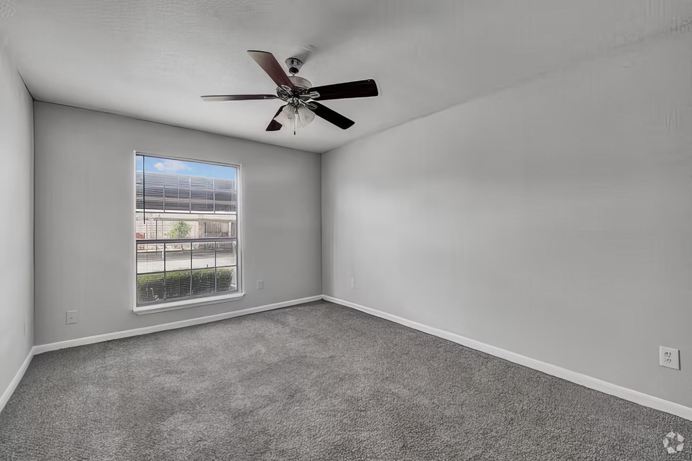 A vacant room with light gray walls and plush carpet. A ceiling fan with dark wooden blades hangs from the ceiling, and a large window provides natural light, revealing an outdoor view. The space is empty and ready for furniture.