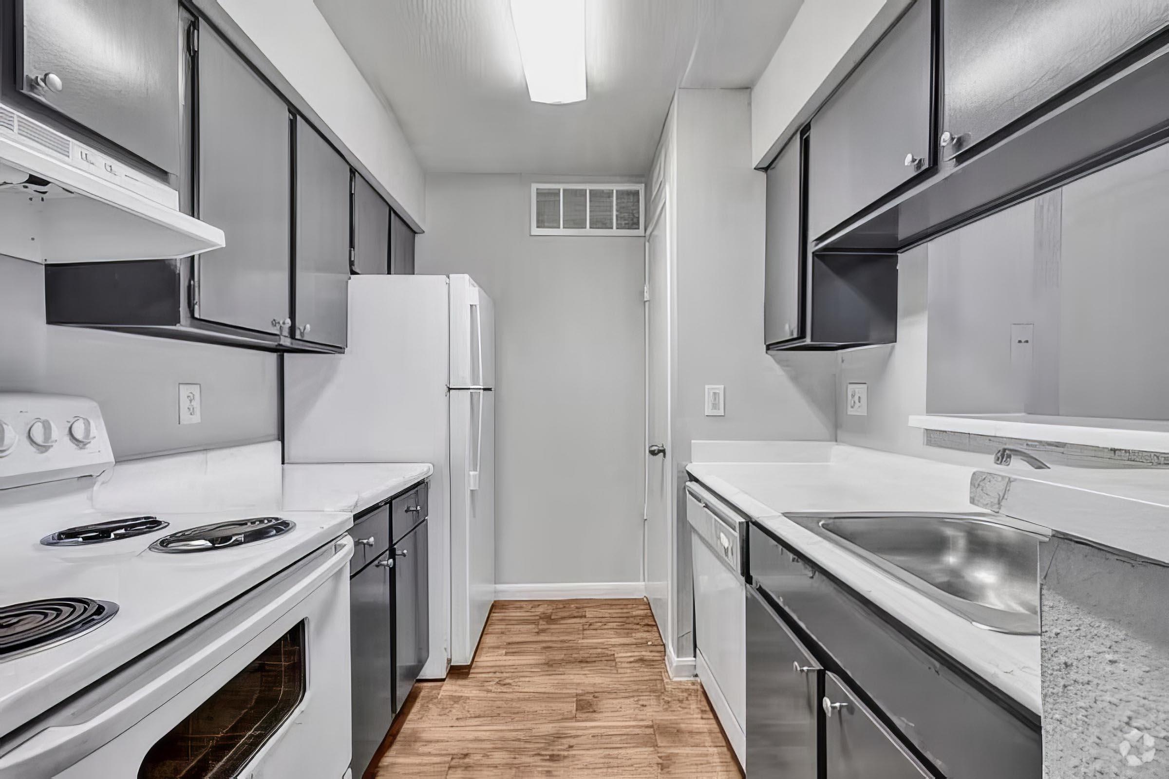A modern kitchen featuring dark gray cabinets, white appliances including a stove and refrigerator, a sink, and light wood flooring. The walls are painted in a light color, and there is ample countertop space, creating a clean and organized look. Natural light comes from a ceiling fixture.