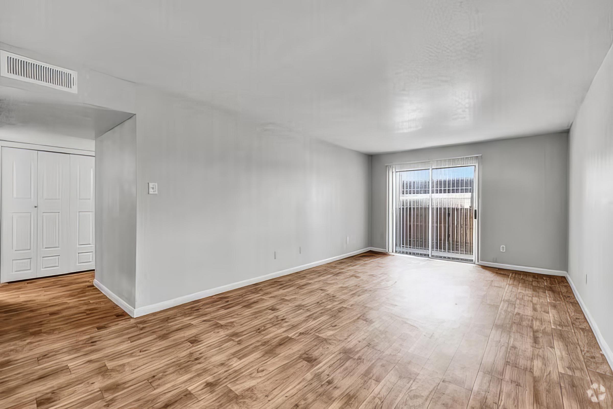 Spacious, well-lit living room featuring light wood flooring and neutral walls. A sliding glass door leads to an outdoor area, while a doorway on the left provides access to a closet. The room is empty, showcasing its potential for furniture arrangement.