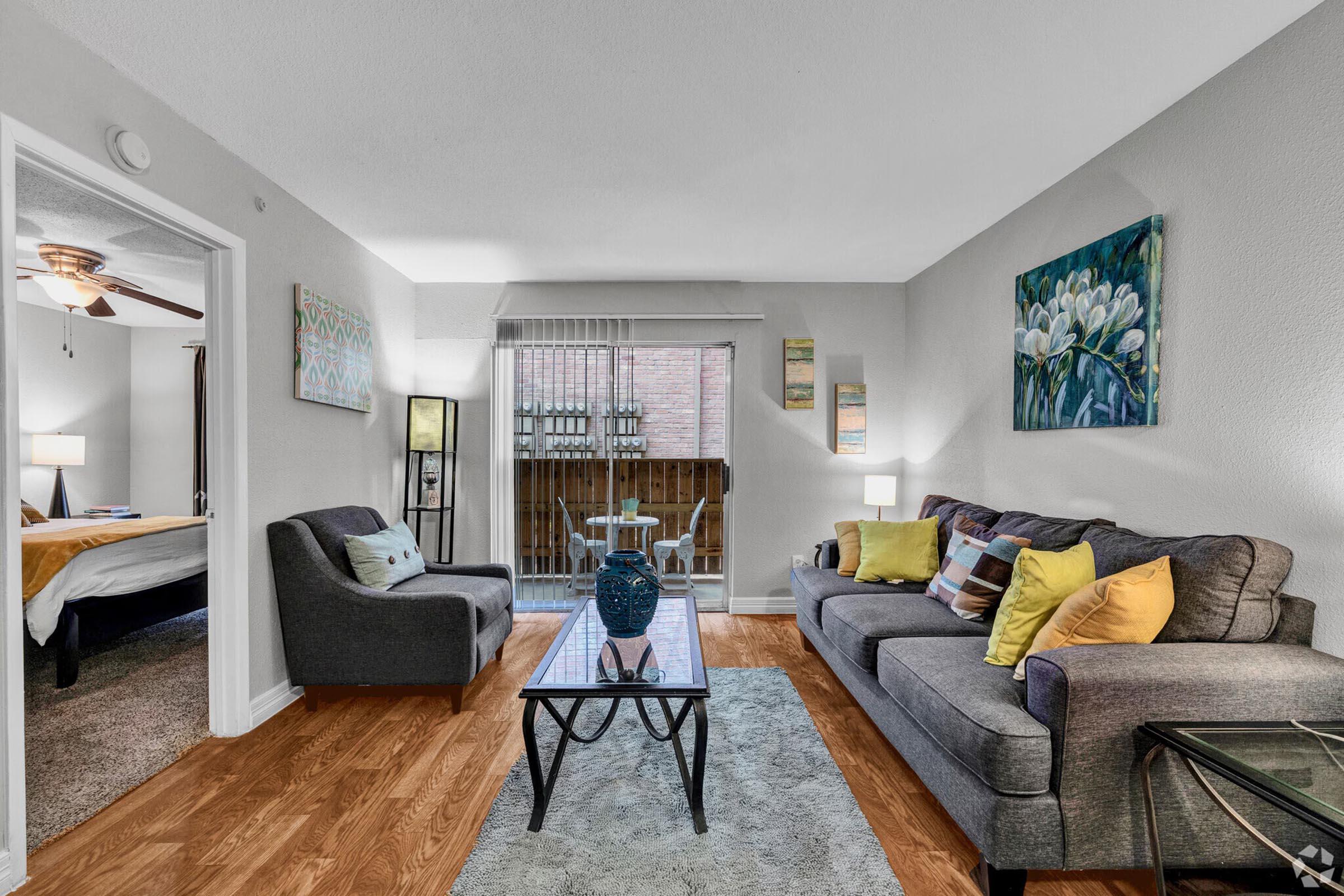 A cozy living room featuring a gray sofa with colorful cushions, a glass coffee table, and a blue decorative vase. There's a comfortable chair and a door leading to a balcony. The room has warm wood flooring and soft lighting, with artwork on the walls adding a touch of color. Visible is a glimpse of a bedroom through the open door.