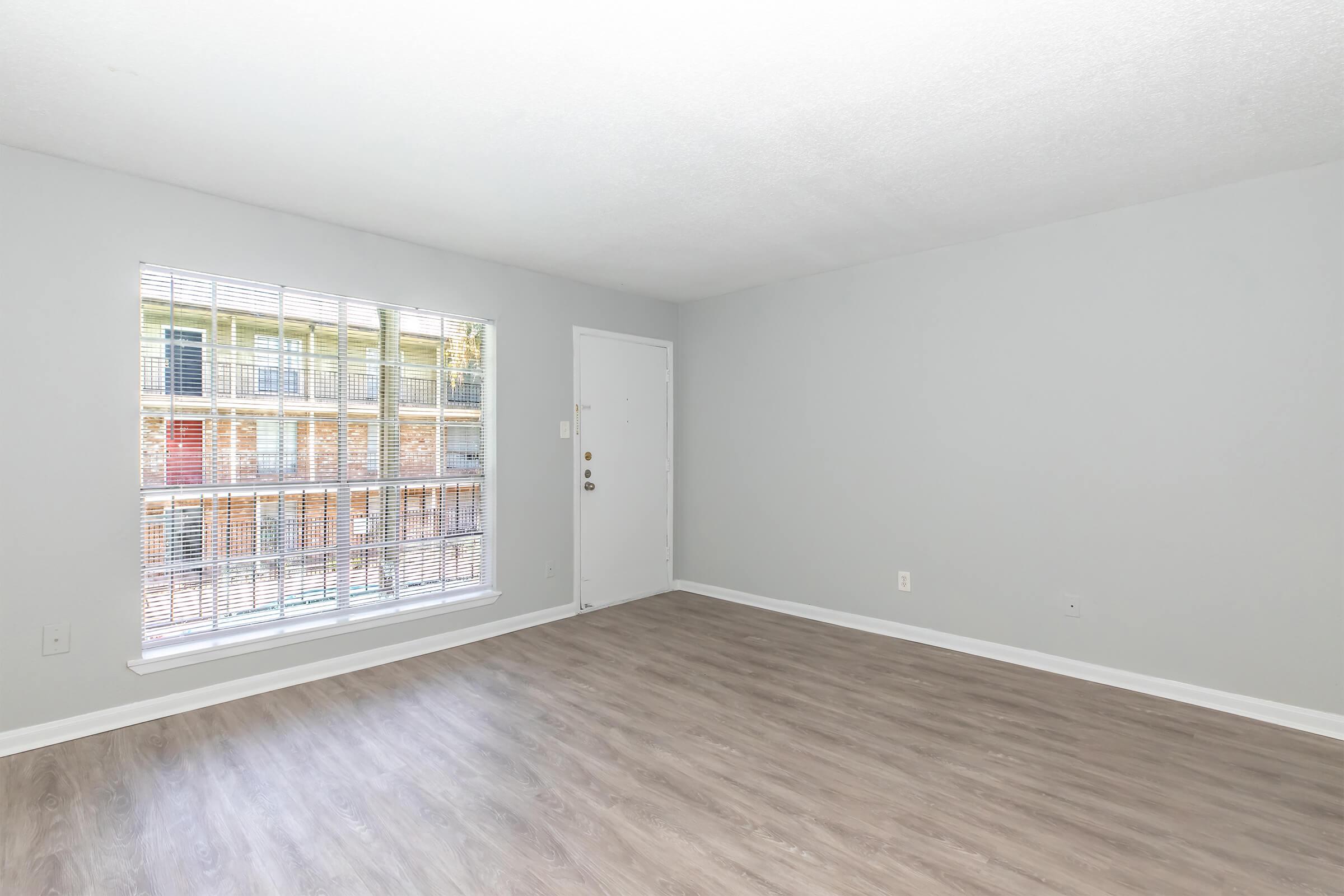 A well-lit, empty living room with light gray walls and a large window featuring white blinds. The floor is a light wood tone, and there is a single door on one wall. The space is clean and has a modern, airy feel, with no furniture present.