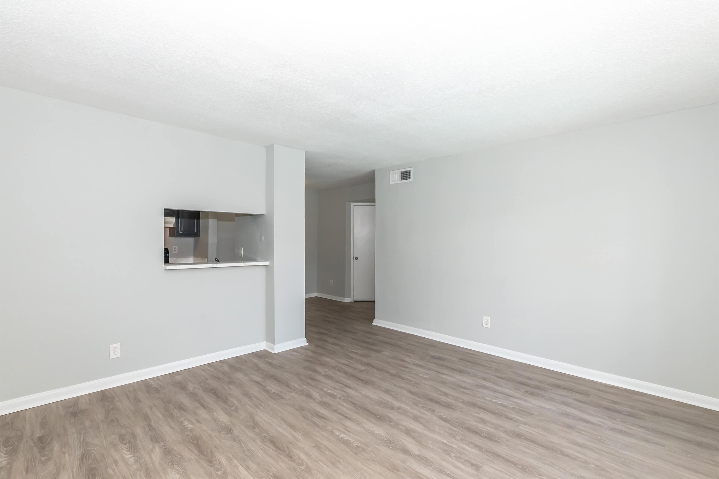 A spacious, empty room with light gray walls and a wood-like floor. A small kitchen area is visible on the left with a bar countertop. The room has no furniture and features a door leading to another area in the background. Natural light brightens the space.