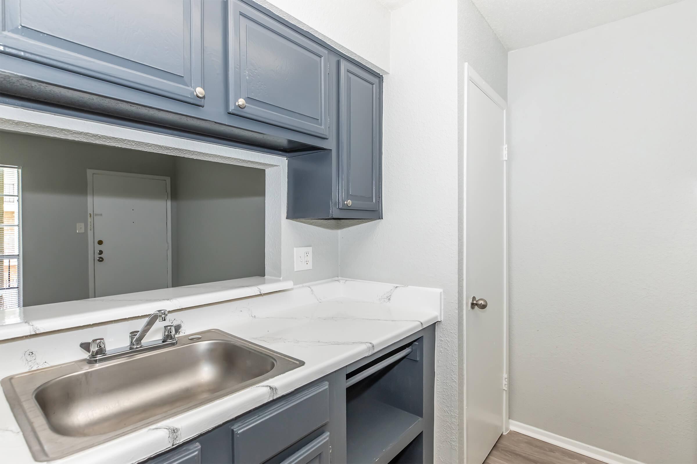 A modern kitchen space featuring dark blue cabinetry and a marble counter with a sink. A small opening leads to another room, and a door on the right suggests access to additional space. The walls are painted light gray, providing a neutral backdrop.
