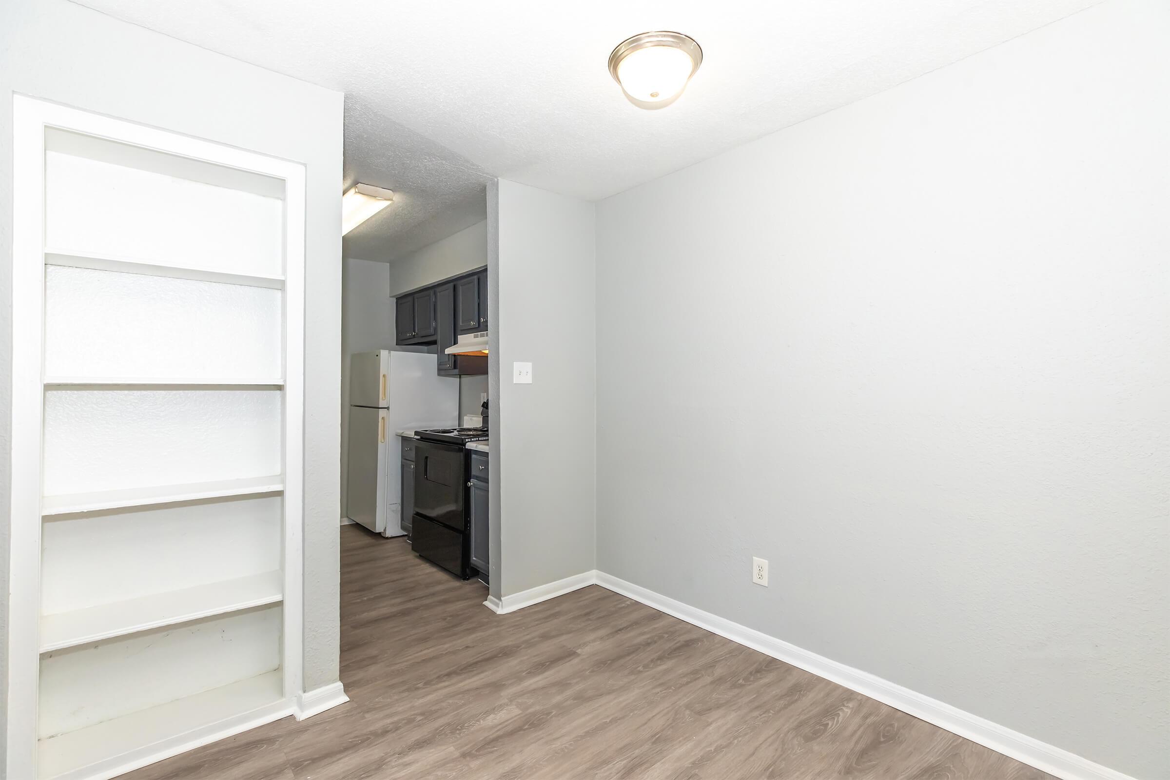 A small, modern kitchen area with gray walls and laminate flooring. There is a built-in white bookshelf on the left, a refrigerator and stove visible in the background, and a ceiling light fixture. The space has a clean and minimalistic appearance.