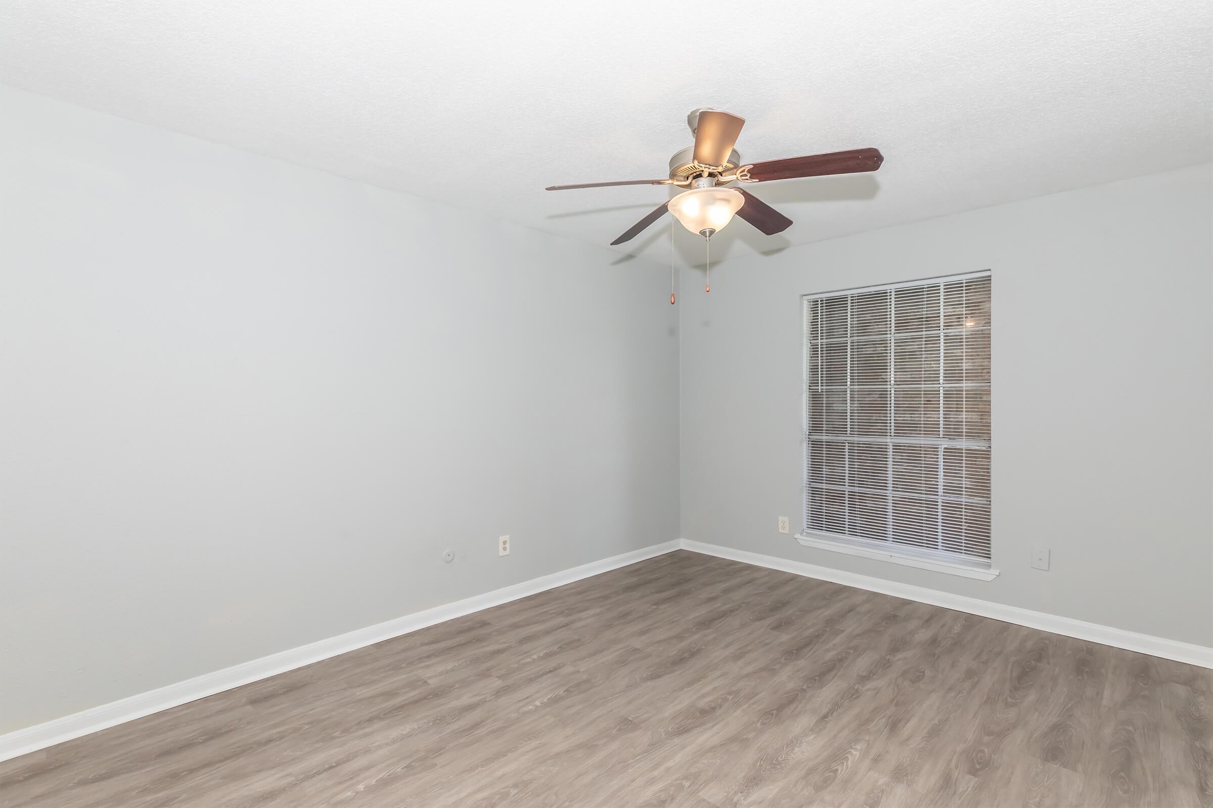 A light gray room with a ceiling fan and a single window featuring horizontal blinds. The flooring is a light wooden style, and the walls are undecorated, creating a simple, clean aesthetic. The space is empty, suggesting it is ready for furniture or decoration.
