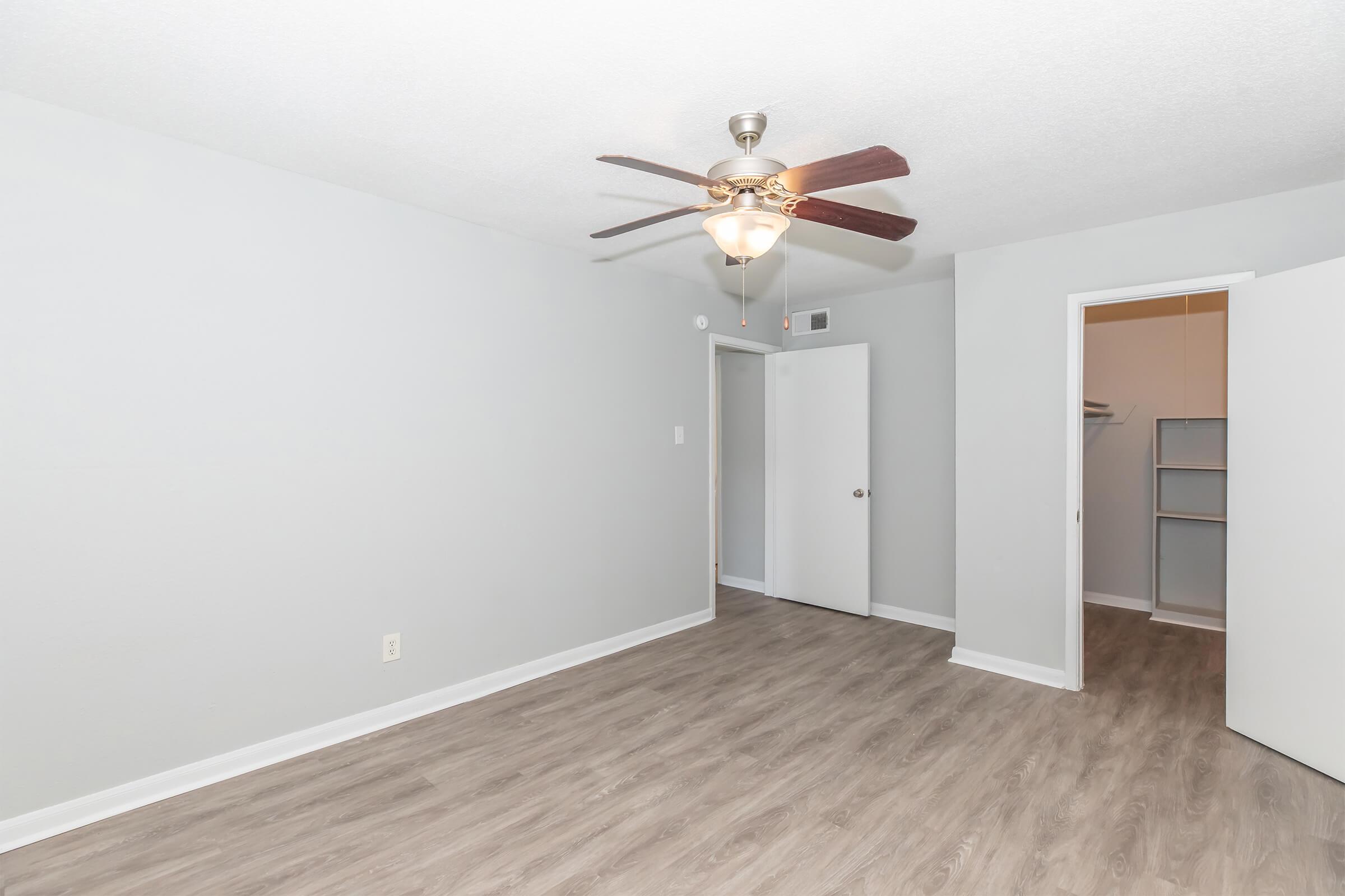 A well-lit room featuring light gray walls and hardwood-style flooring. The ceiling has a ceiling fan with wooden blades. There are two doors: one leads to a closet, and the other is an entryway. The space is empty, providing a blank canvas for decorating.