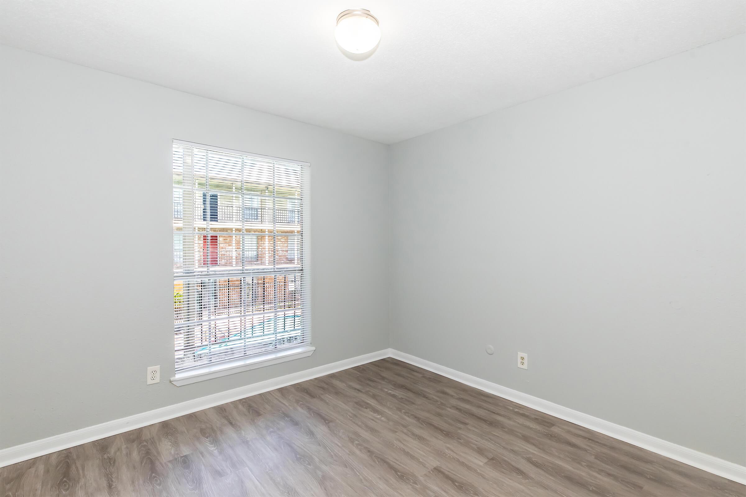 Empty room with light gray walls and a wood-look floor. A window on the left side allows natural light to enter, featuring white blinds. The ceiling has a simple light fixture, and there are no furnishings or decorations visible in the space.