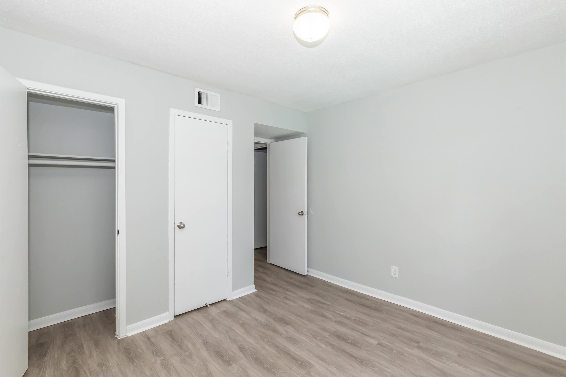 A neutral-colored bedroom with light gray walls and a wood-like floor. There are two doors: one leads to a closet, while the other opens to another room. A ceiling light fixture provides illumination. The space is empty, creating a clean and spacious appearance.