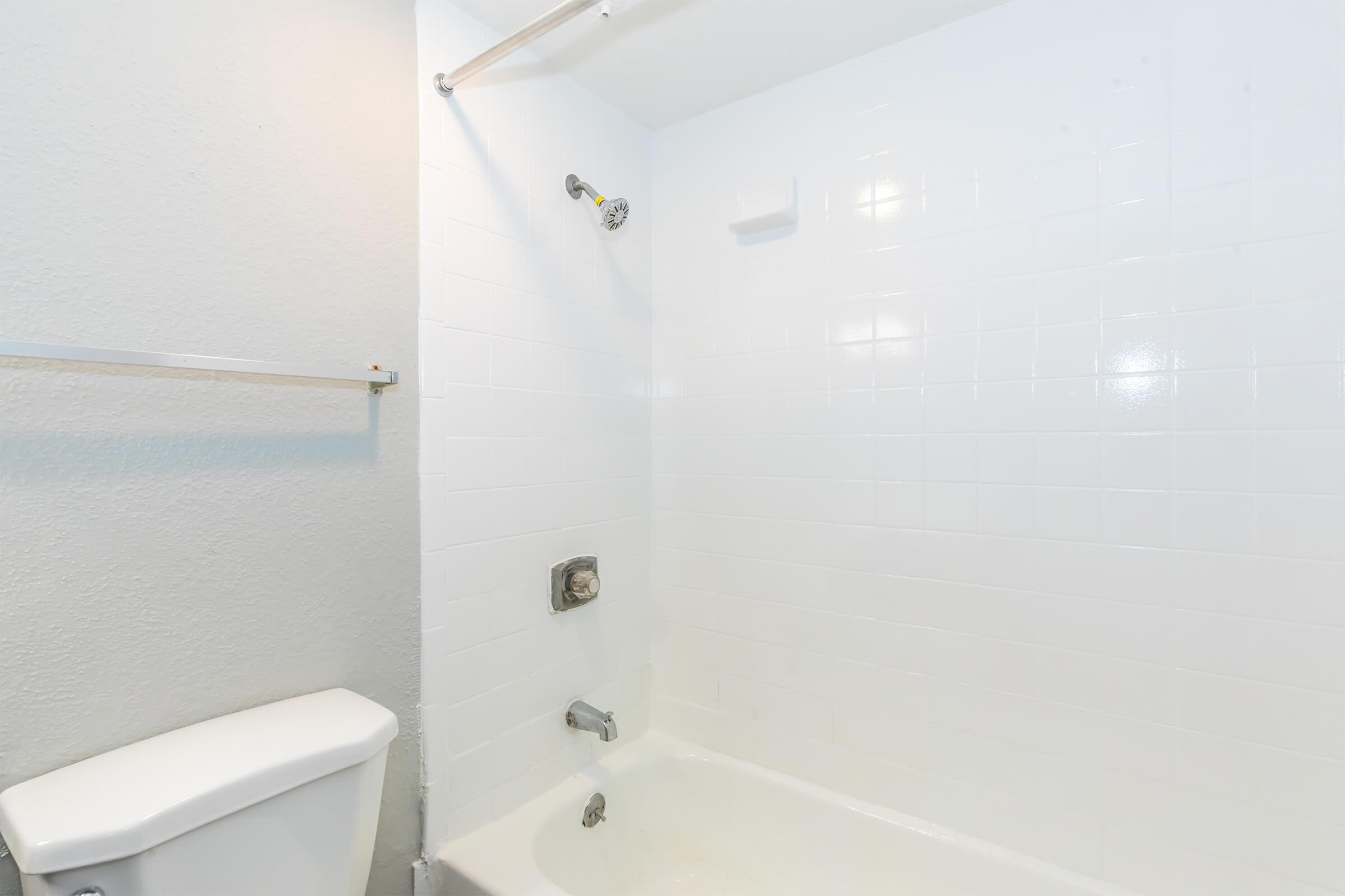 A clean and minimalistic bathroom featuring a white bathtub, a shower head mounted on the wall, and a toilet. The walls are tiled in white, and there is a towel bar present. The overall appearance is bright and well-maintained.