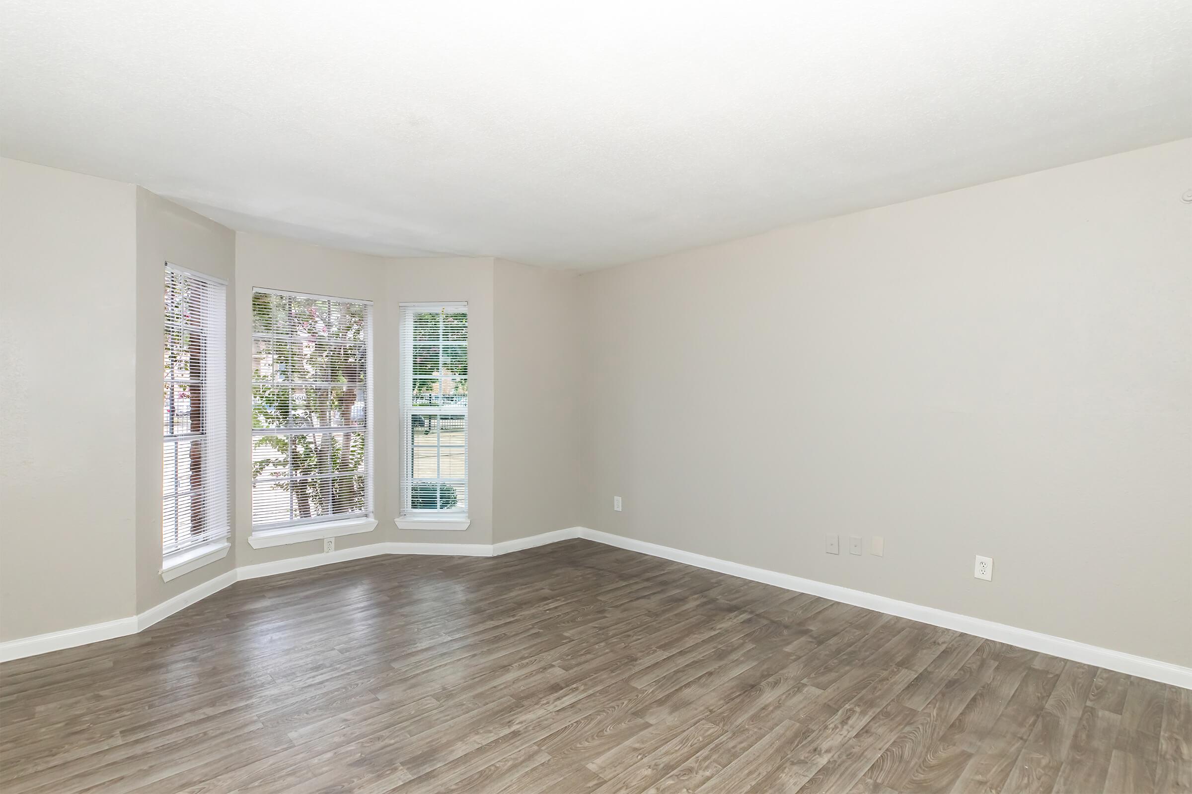 A spacious, empty living room featuring light gray walls, large windows with white blinds, and a laminate wood floor. The room has a clean and bright appearance, with natural light coming through the windows. It is designed for versatility in furnishings and decor.