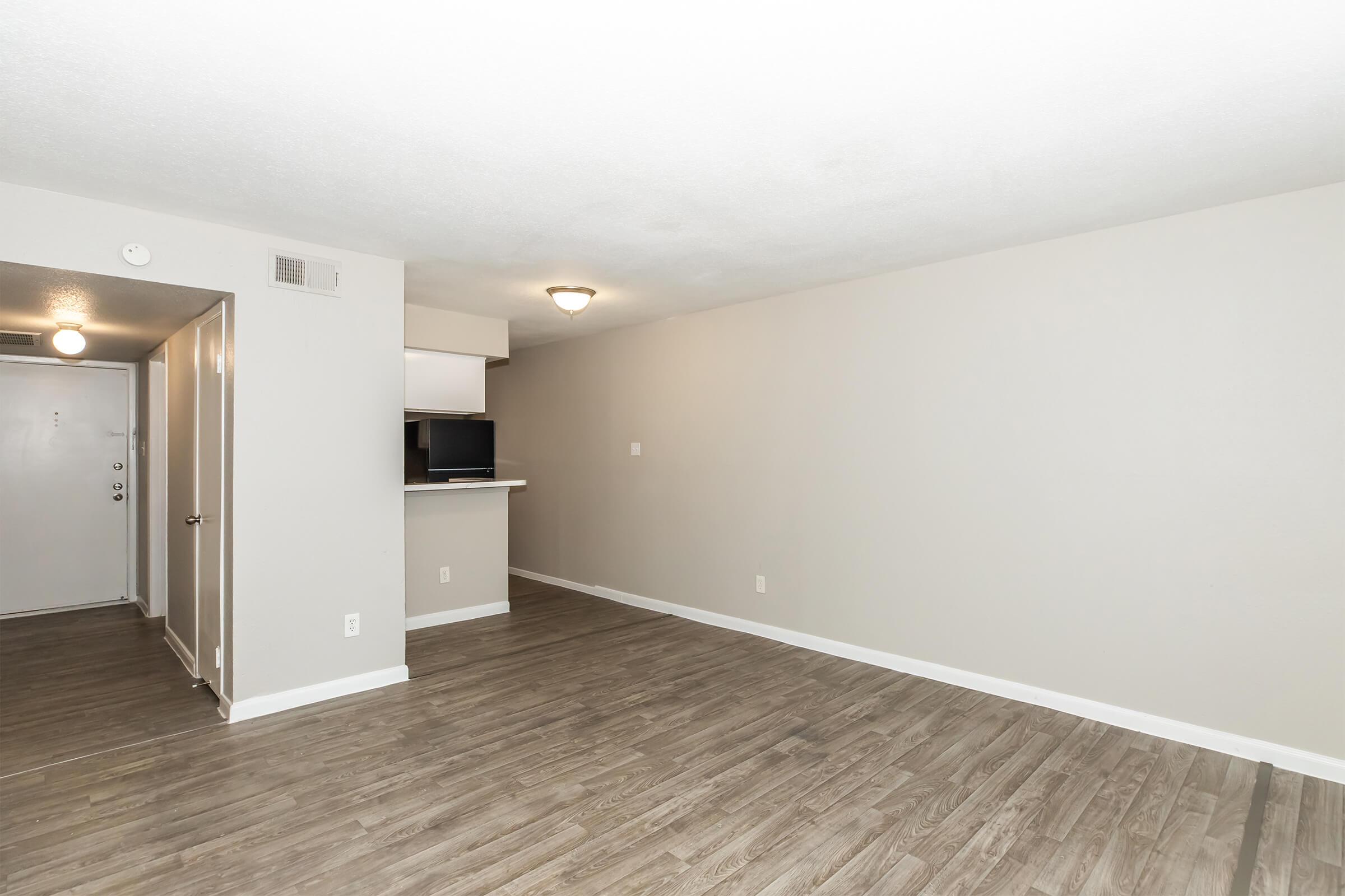 A spacious, empty apartment living area with light-colored walls and wood-like flooring. The room features a ceiling light and a small bar area adjoining an unseen kitchen. There is a door leading to the entrance on the left, adding to the open feel of the space.