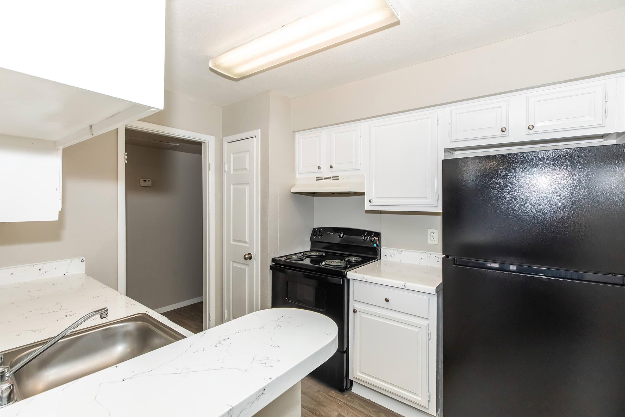 A modern kitchen featuring white cabinets, a black stove and refrigerator, and a sink with a marble countertop. The walls are painted in neutral tones, and there is good lighting from a ceiling fixture. A doorway leads to another room, indicating an open layout.