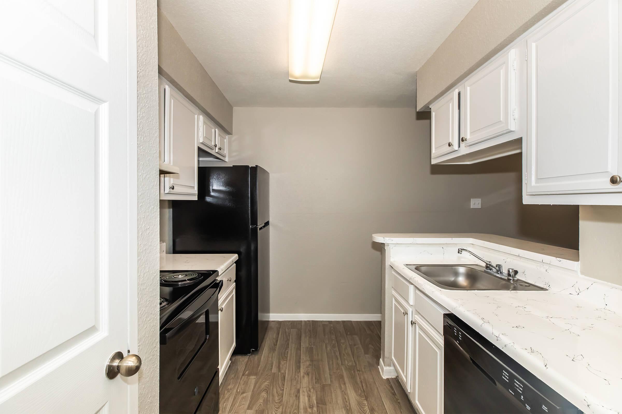 A modern, compact kitchen featuring white cabinetry, black appliances including a refrigerator and stove, and a granite-style countertop. The kitchen has laminate flooring and neutral-colored walls, providing a clean and inviting atmosphere. Well-lit with an overhead light fixture.