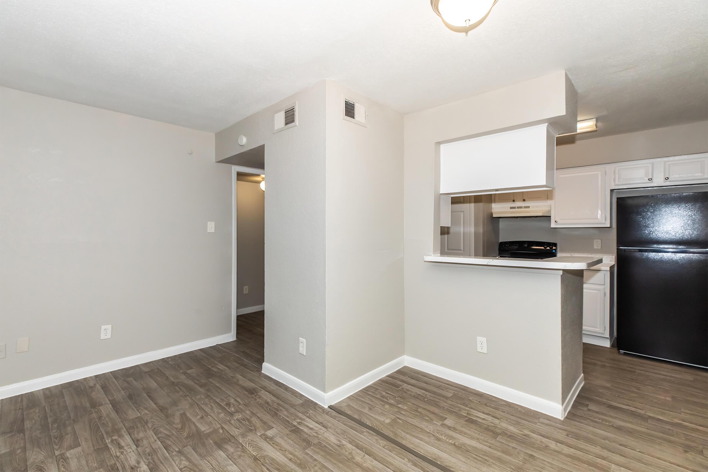 An interior view of a modern apartment featuring open living space, light gray walls, and wood-style flooring. A kitchenette with white cabinetry and a black refrigerator is visible in the background. The layout suggests a cozy and inviting atmosphere.