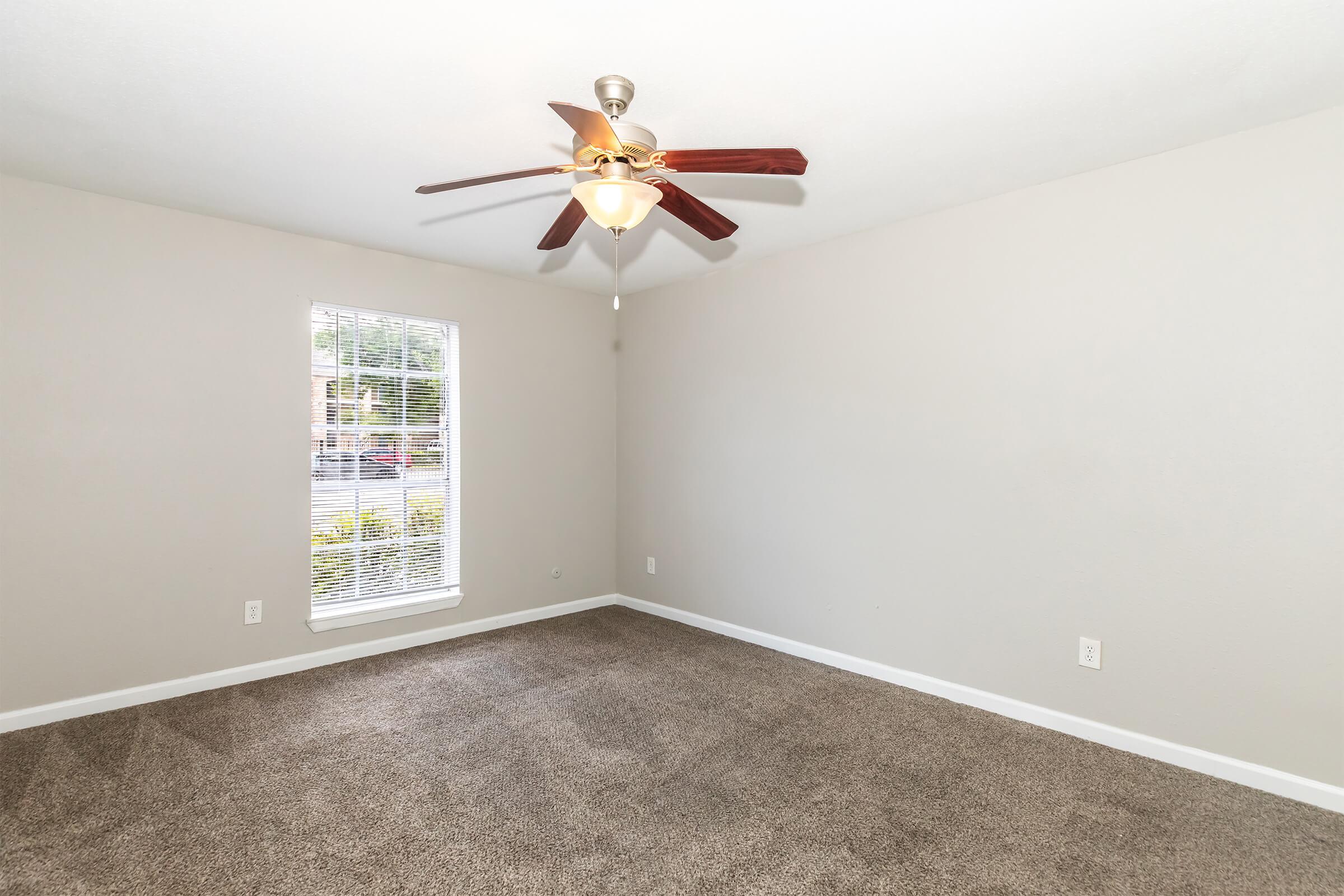 A well-lit room featuring a ceiling fan with wooden blades, beige walls, and a large window allowing natural light. The floor is covered with soft, light-colored carpeting. The space is empty, providing a blank canvas for decoration.