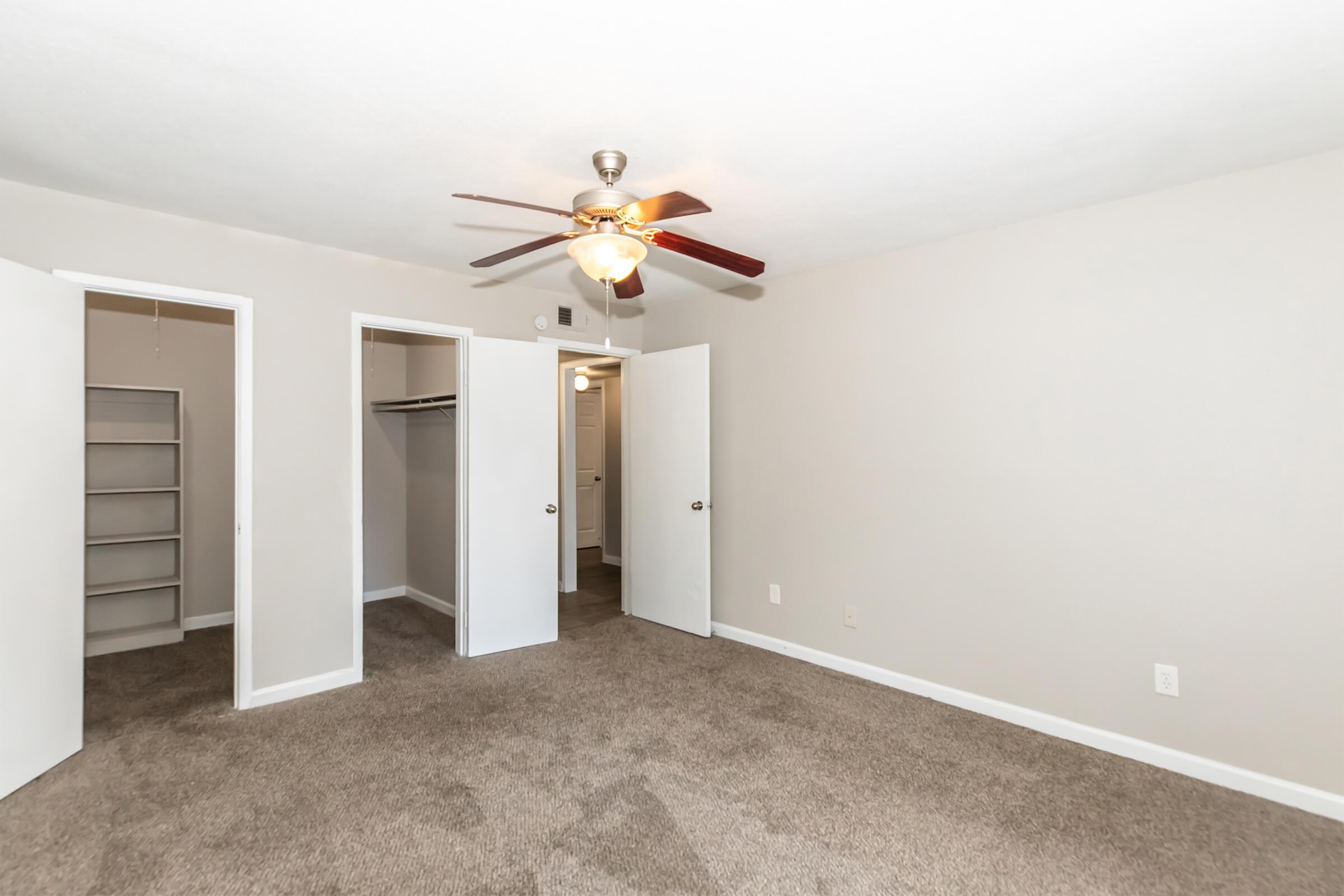 A spacious room featuring beige walls and carpet flooring, complete with a ceiling fan. There are two open white doors leading to closets. A third door is seen in the background, suggesting access to a bathroom or another room. The lighting is bright and inviting, creating a cozy atmosphere.