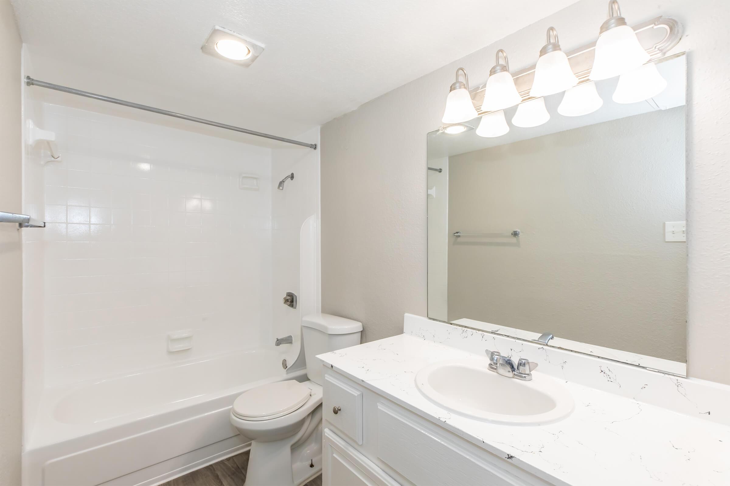 A clean and bright bathroom featuring a white bathtub with a shower, a toilet, and a vanity with a sink. There is a large mirror above the sink and five light fixtures illuminating the space. The walls are a light color, and the flooring appears to be a light wood or laminate.