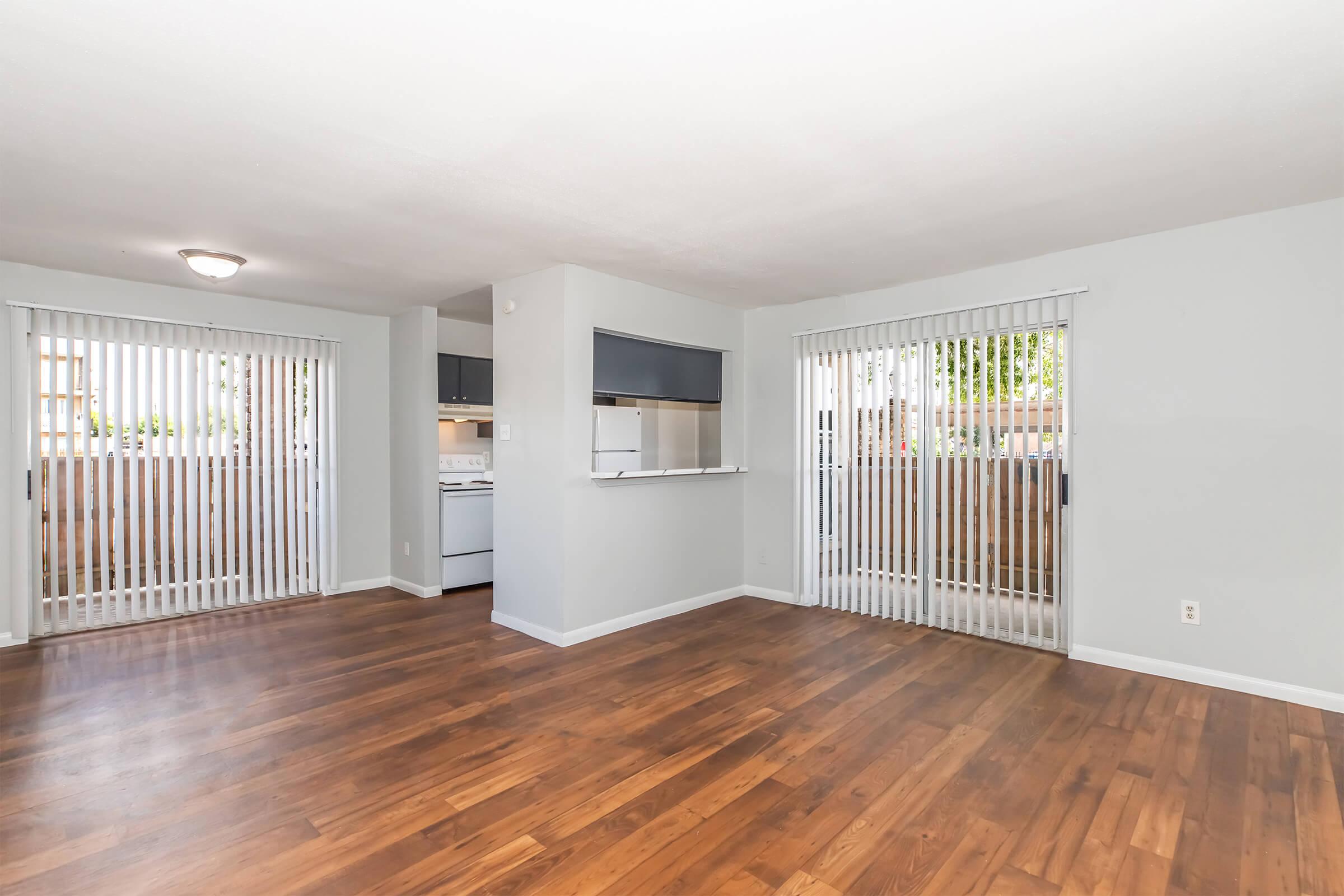 An empty living room featuring hardwood flooring, light gray walls, and vertical blinds on large windows. There is an open kitchen area visible, with a white stove and a countertop separating it from the living space. Natural light is flowing in through the windows, creating a bright atmosphere.