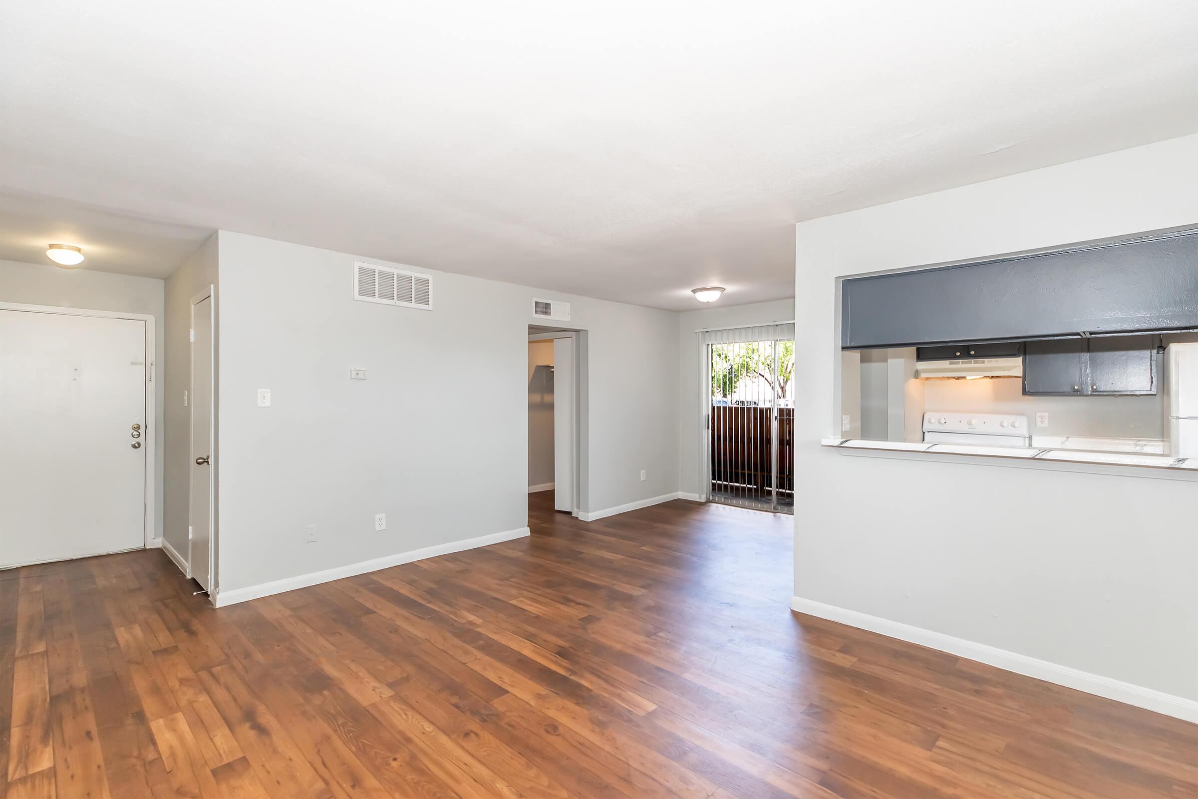 Interior view of a modern, spacious living area featuring light gray walls, hardwood flooring, and an open layout. The room includes a doorway leading to another space, and a kitchen area visible in the background with a counter. Natural light comes through windows, enhancing the bright atmosphere.