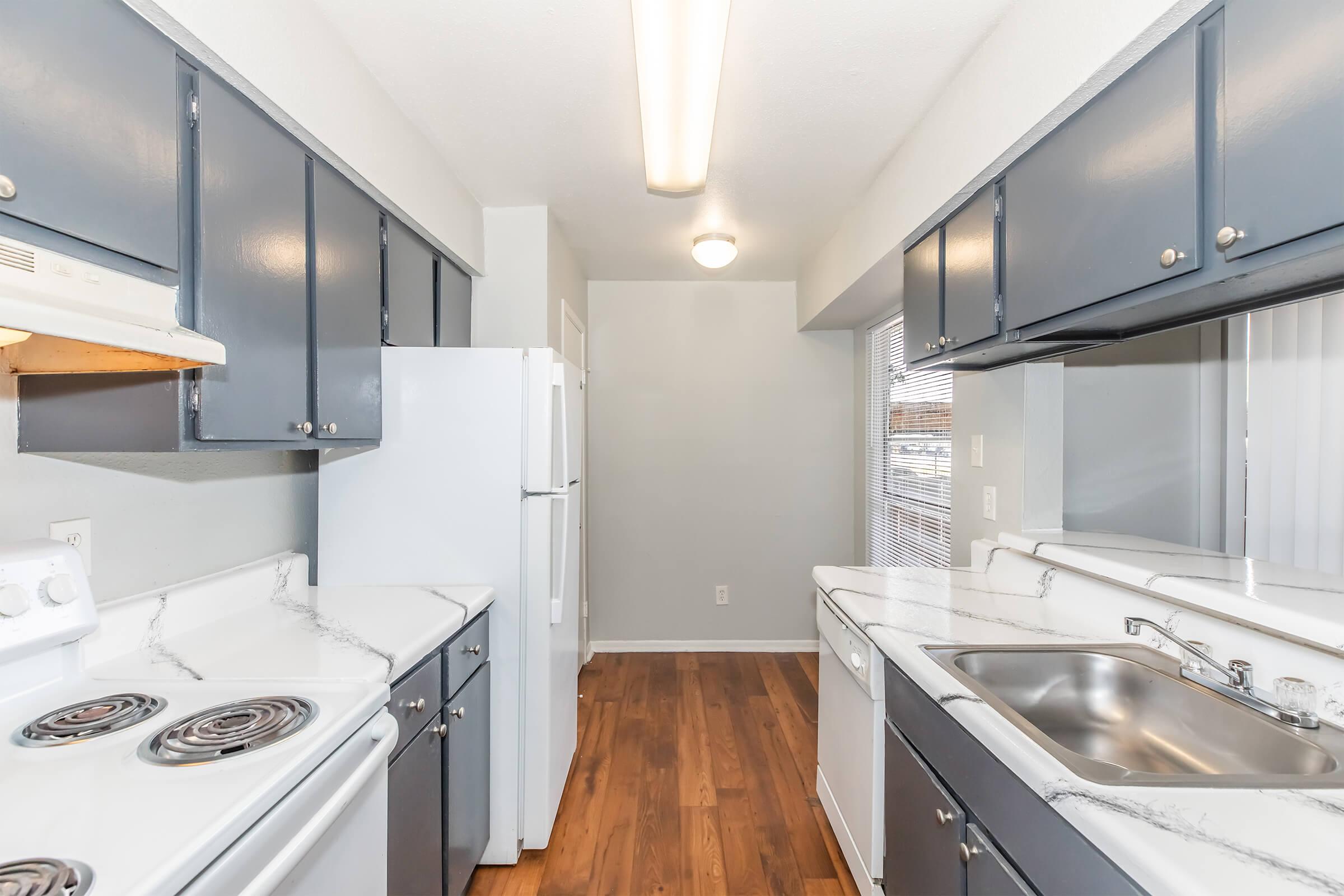 Modern kitchen with gray cabinets, a white refrigerator, a stove with a black cooktop, and a white sink. The countertops have a marble finish, and there is natural light coming in from a nearby window, illuminating the space. The flooring is wooden, creating a warm atmosphere.