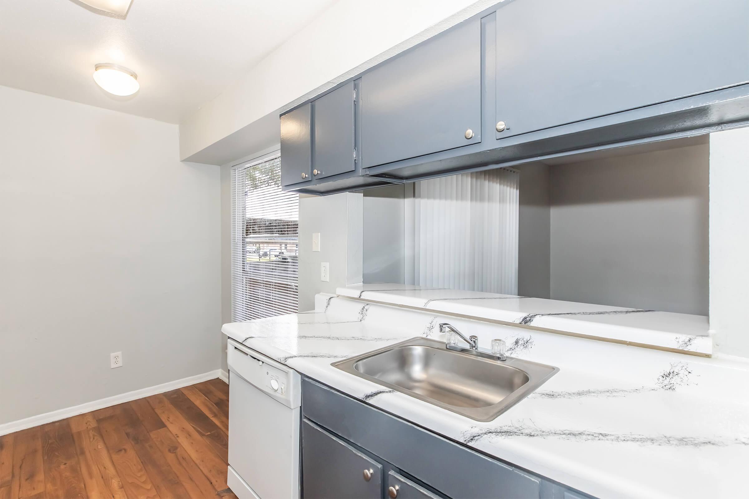 Modern kitchen featuring dark blue cabinetry, a stainless steel sink, and a white marble countertop. The floor is made of light wood planks. A window provides natural light, and there is a dishwasher visible next to the sink. The walls are painted in a light gray color, creating a bright and airy feel.