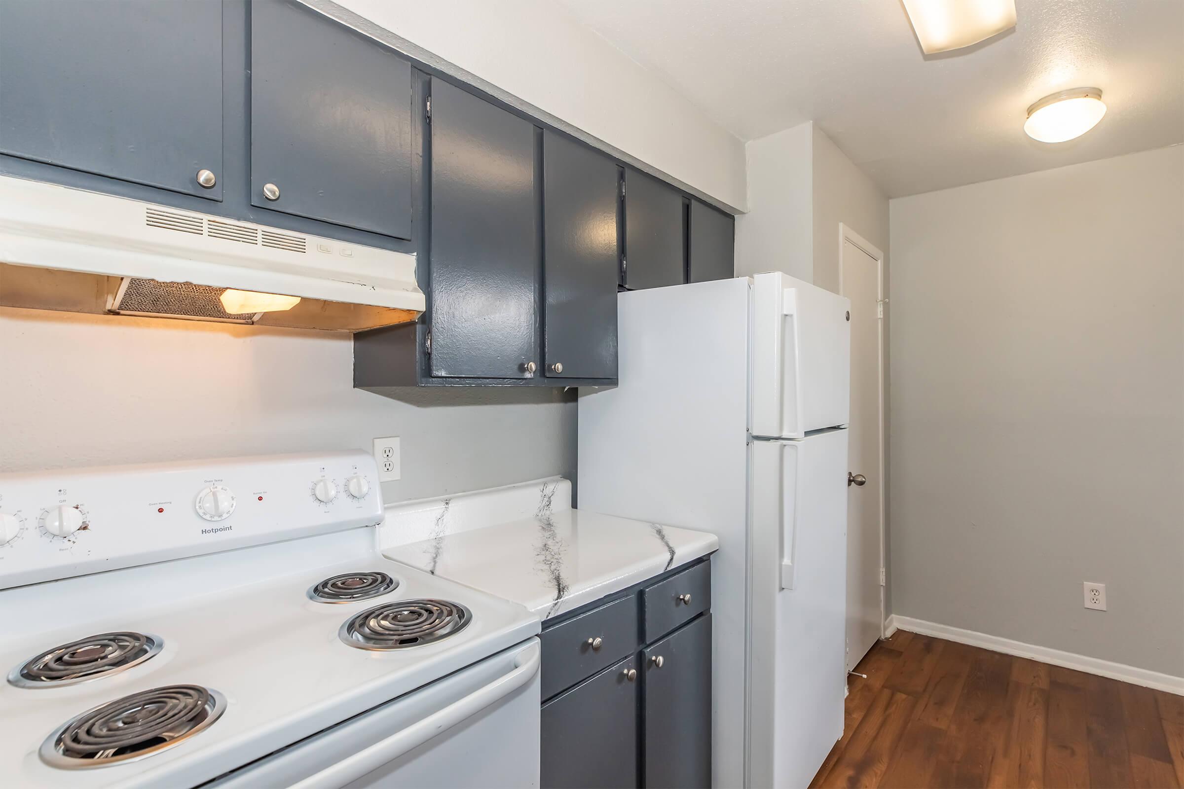 A modern kitchen featuring a white stove with four burners, a white refrigerator, dark gray cabinets, and a light-colored countertop. The walls are painted in a neutral shade, and the floor has wood-like laminate. The space is well-lit with overhead lighting.