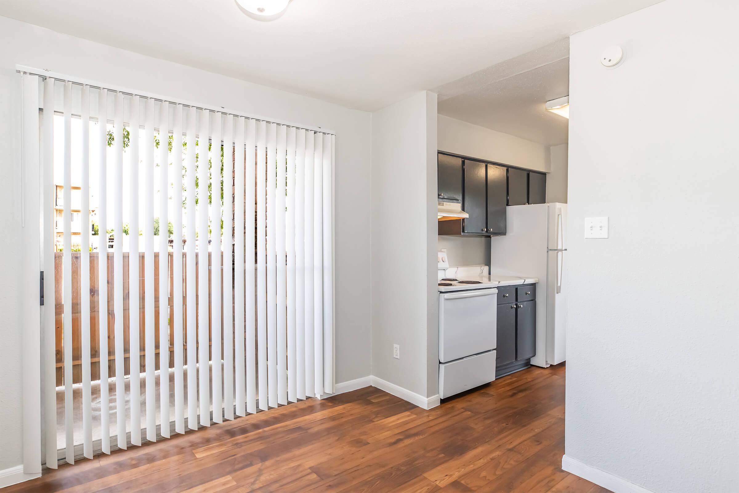 A bright kitchen area featuring gray cabinets, a white fridge, and a stove. There's a white sliding door with vertical blinds leading to a balcony, and warm wooden flooring throughout. The walls are painted light gray, creating a spacious and inviting atmosphere.