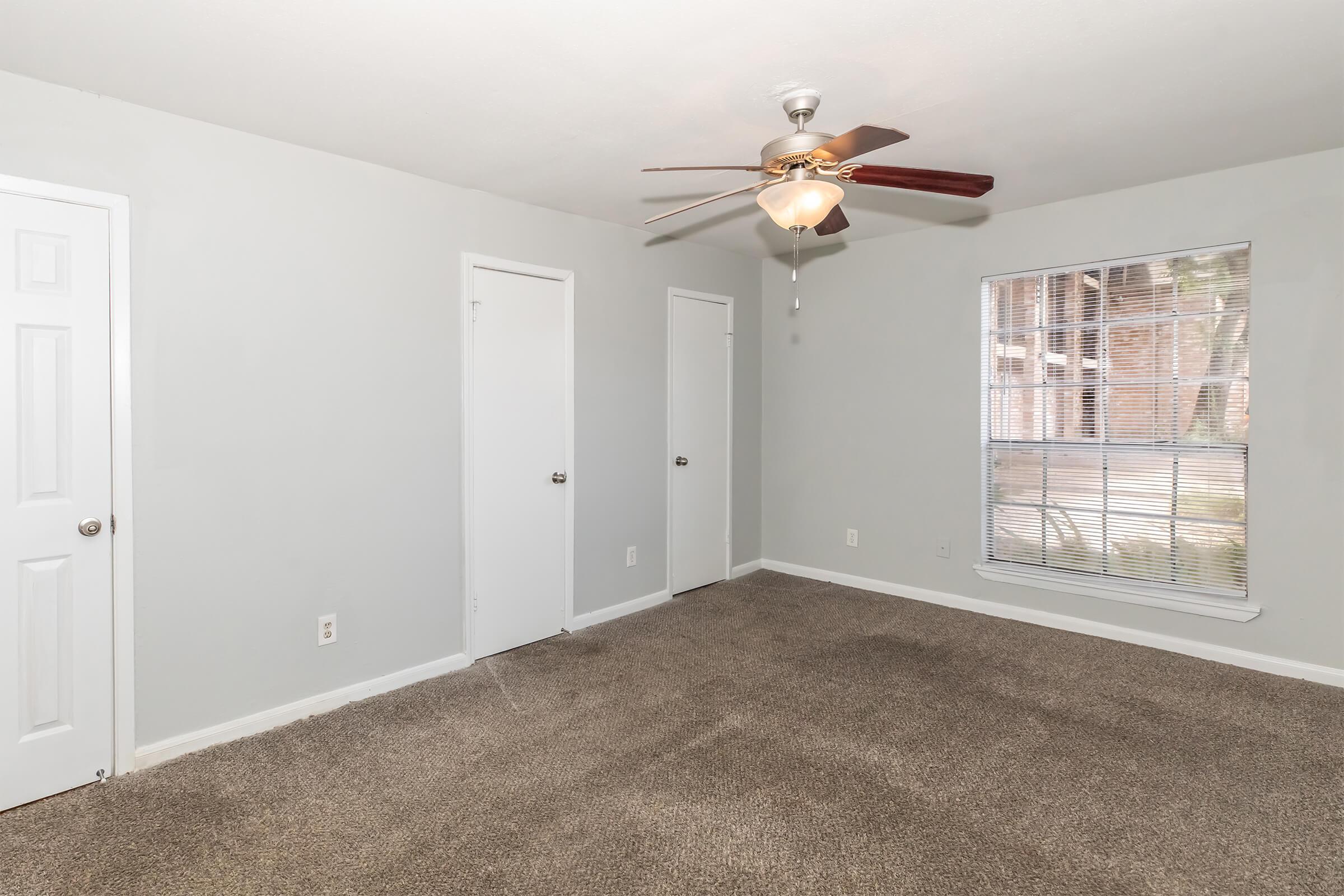Empty room featuring light gray walls, a ceiling fan with wooden blades, and plush brown carpeting. There are two closed white doors on the left side and a window with blinds on the right, allowing natural light to fill the space. The room is clean and well-lit, creating a welcoming atmosphere.
