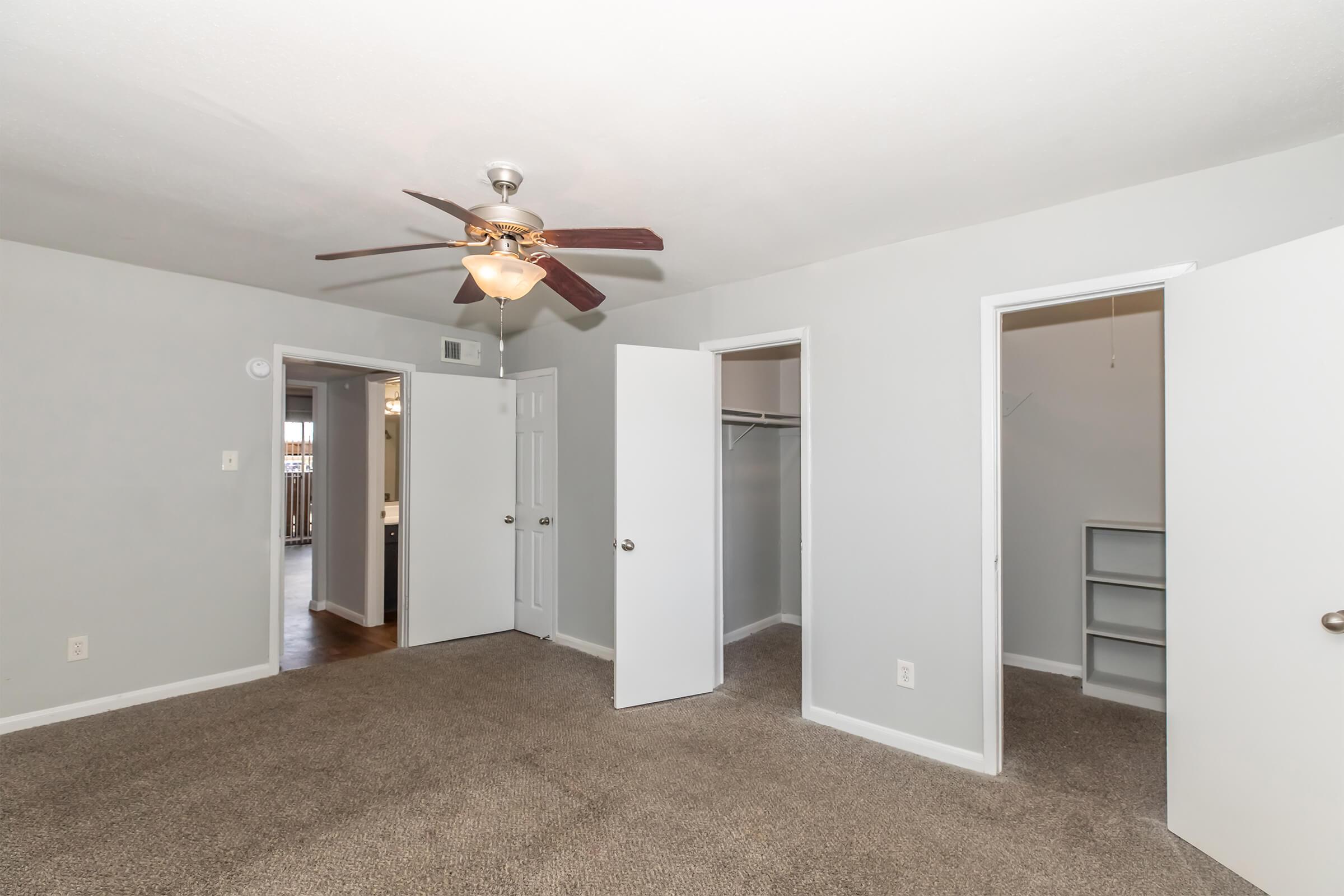 A well-lit bedroom featuring beige carpeting, a ceiling fan with light fixture, and two open doors leading to closets. The walls are painted in a light gray color, enhancing the spacious feel of the room. A doorway in the background leads to another room, adding to the open layout of the space.