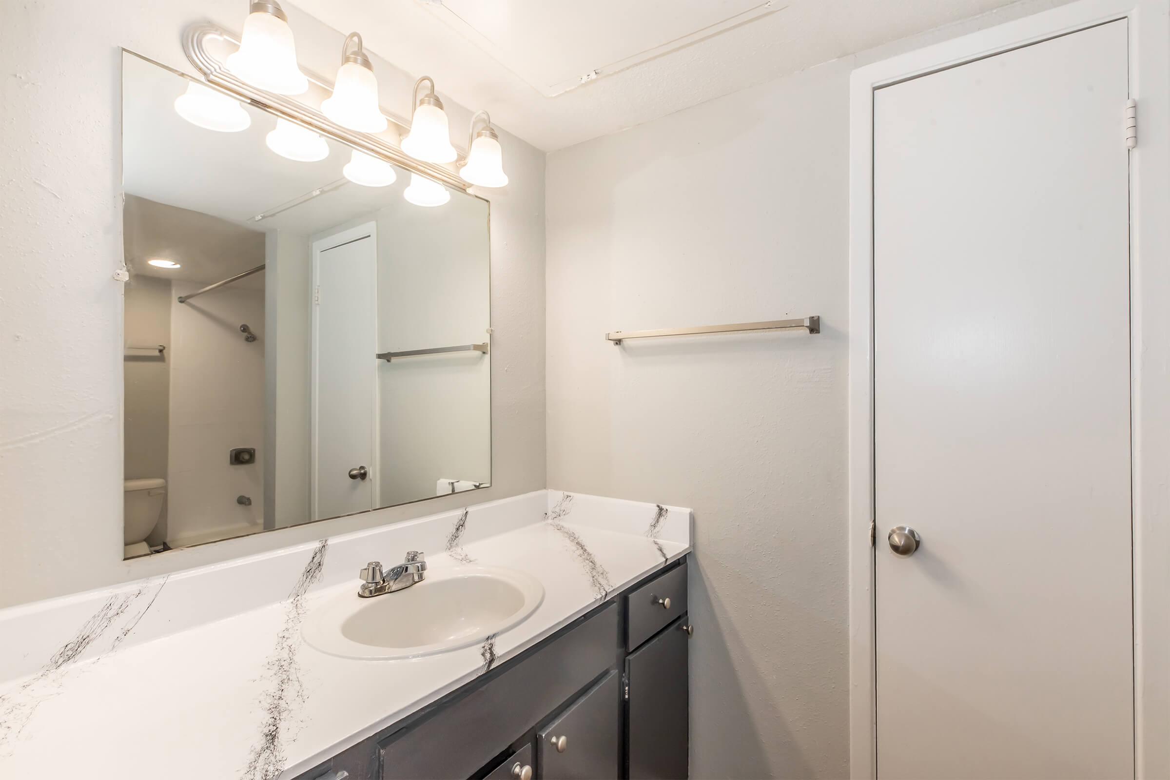 A clean and modern bathroom featuring a white countertop with a sink, a wall-mounted mirror with bright lighting above, and a door. The walls are painted a light color, and there is a towel bar to the side. The decor is minimalistic, creating a spacious feel.