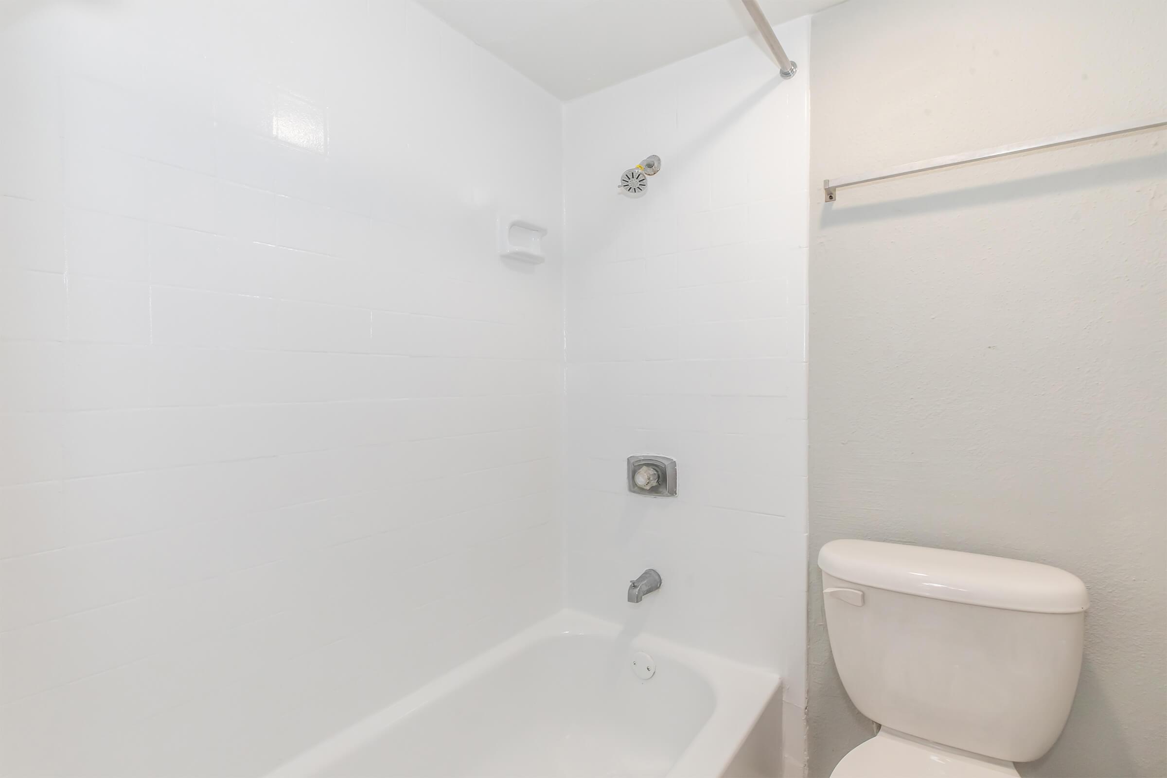 A clean, modern bathroom with a white bathtub, shower fixture, and a toilet. The walls are plain white tiles, creating a bright and spacious atmosphere. There is a towel rack mounted on the wall, enhancing the minimalistic design.