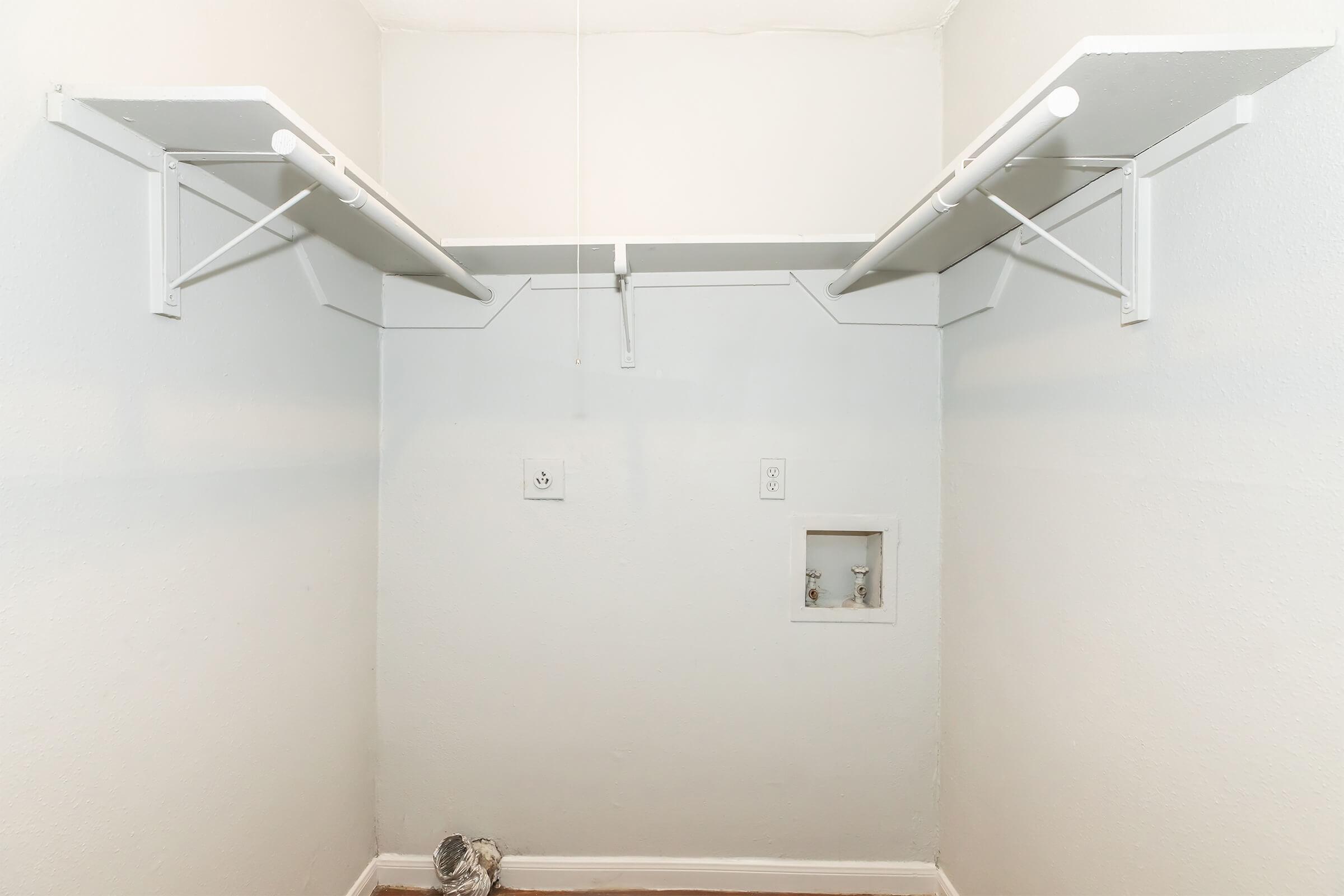 A simple, empty closet with two white shelves mounted on the walls. There are electrical outlets and plumbing fixtures visible on the wall, and the floor is bare. The walls are painted light gray, creating a clean and minimalistic appearance.